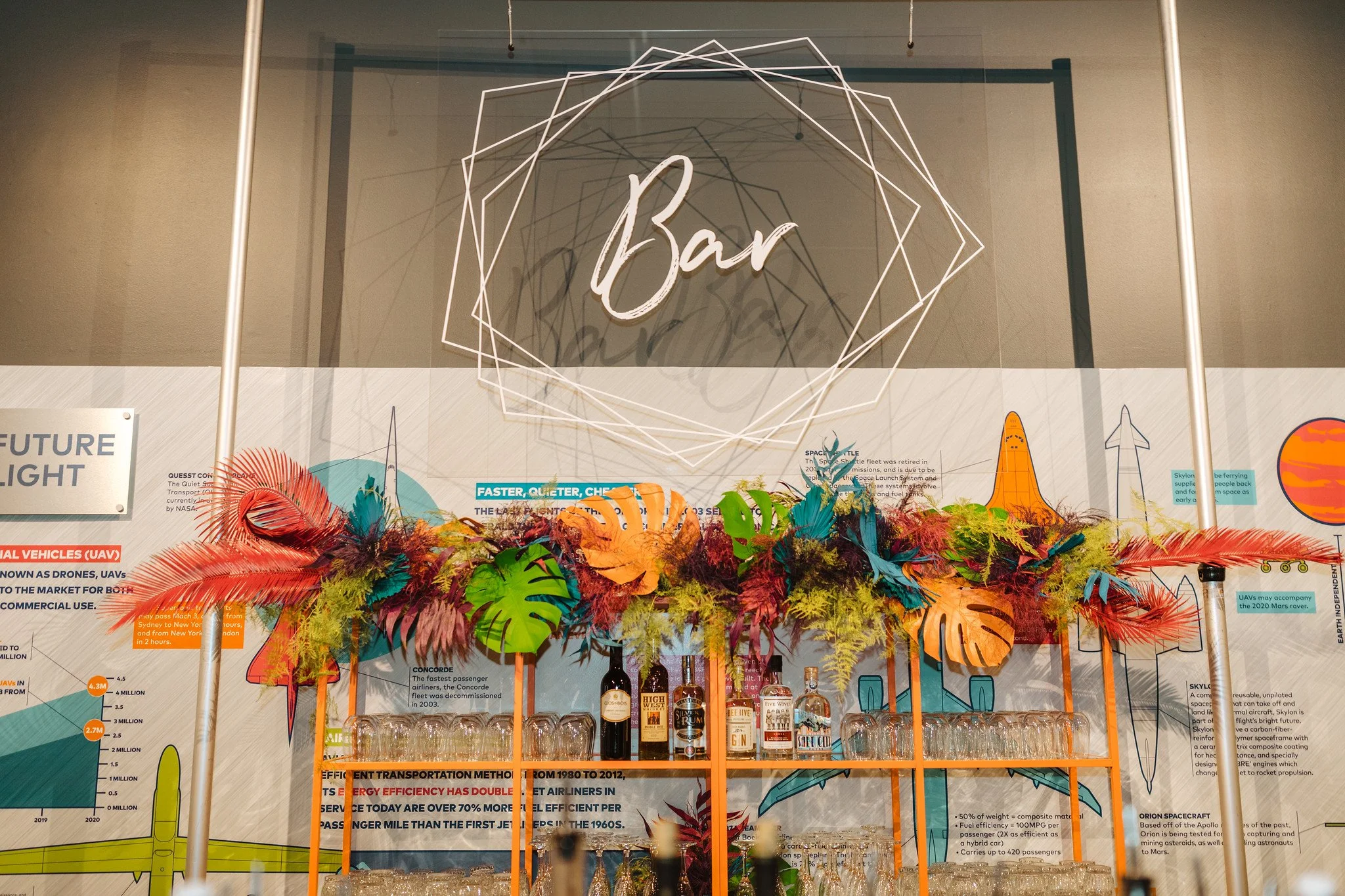A bar with colorful tropical decorations, including red, orange, blue, and purple feathers and leaves, and a variety of liquor bottles. Behind the bar are informational posters about space exploration and transportation methods.