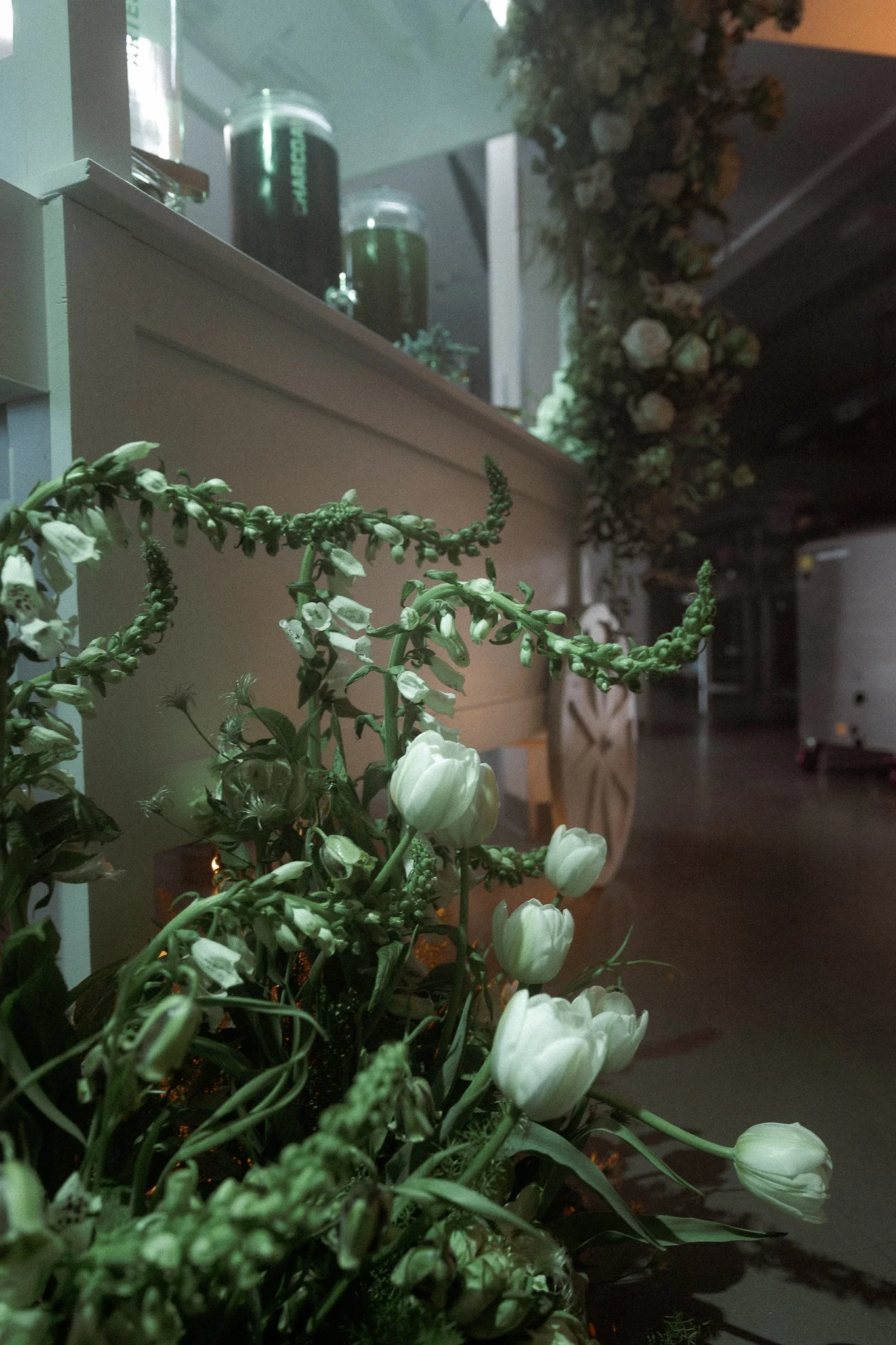 Indoor scene with white tulips and green foliage in a glass vase in the foreground, a wooden table and chairs, and a floral garland hanging along the wall in the background.
