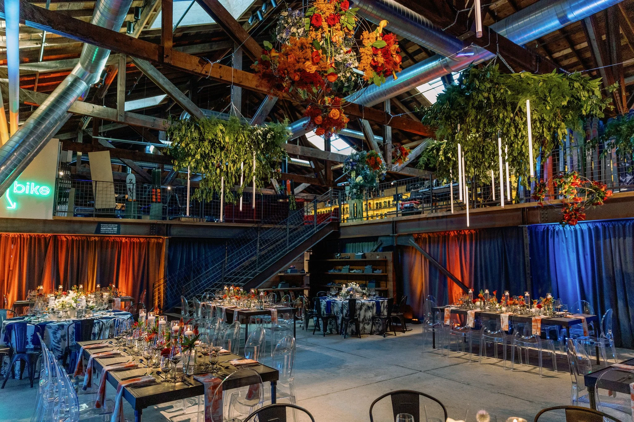 An indoor event space decorated for a celebration, featuring arranged tables with glassware, flowers, and candles, a floral arrangement hanging from the ceiling, visible metal and wood structural elements, and colorful drapery along the lower wall.