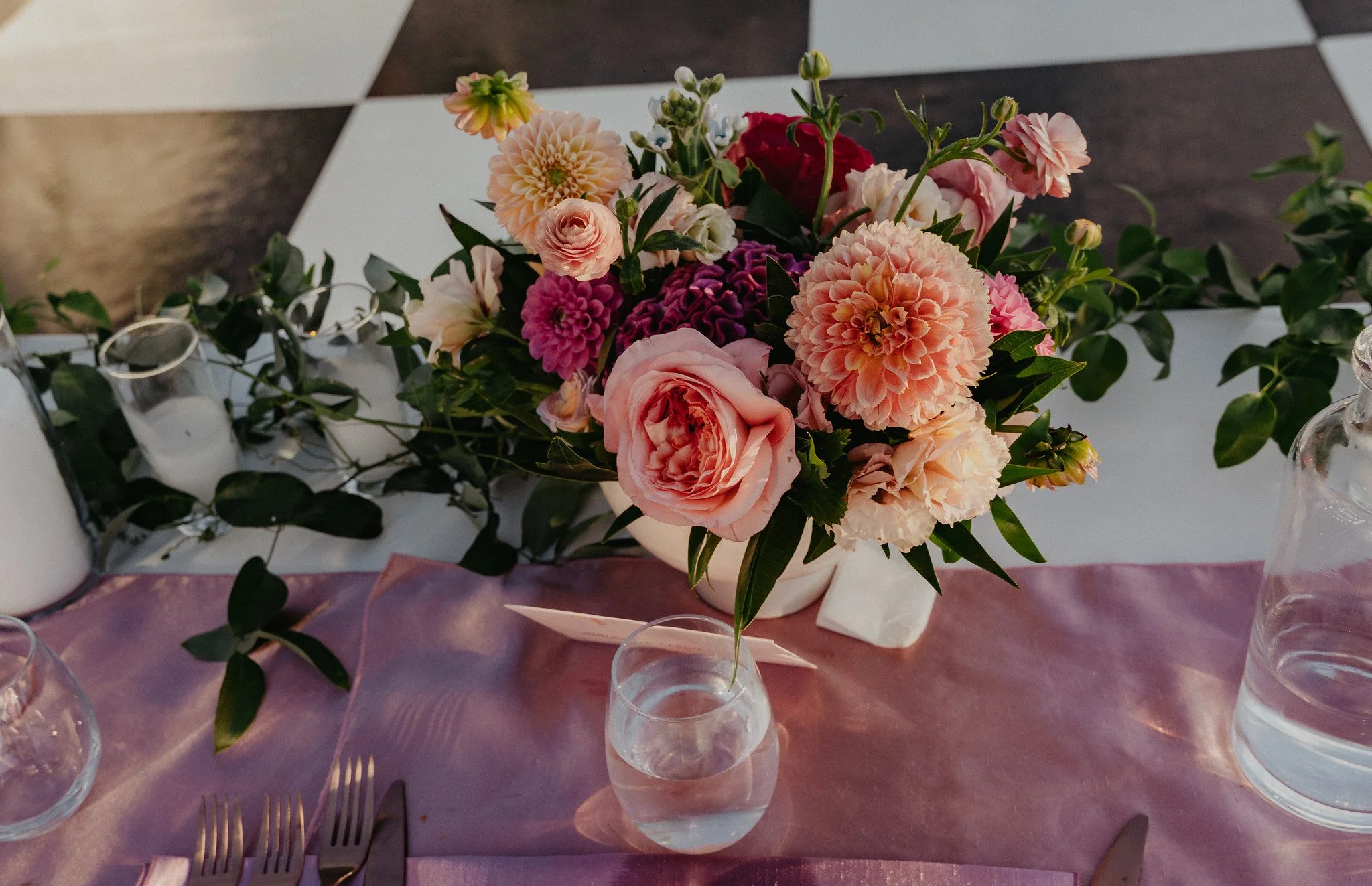 A floral centerpiece with pink, peach, and red flowers on a table with purple tablecloth, surrounded by clear drinking glasses and silver forks.