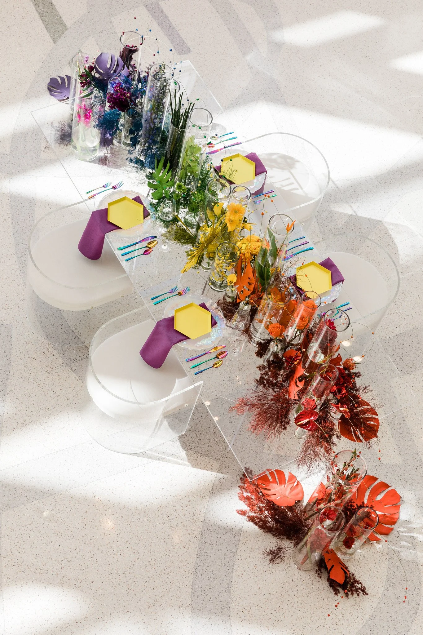 A long table decorated with colorful flowers, glass vases, purple napkins, and multicolored cutlery, set for a celebration or event.
