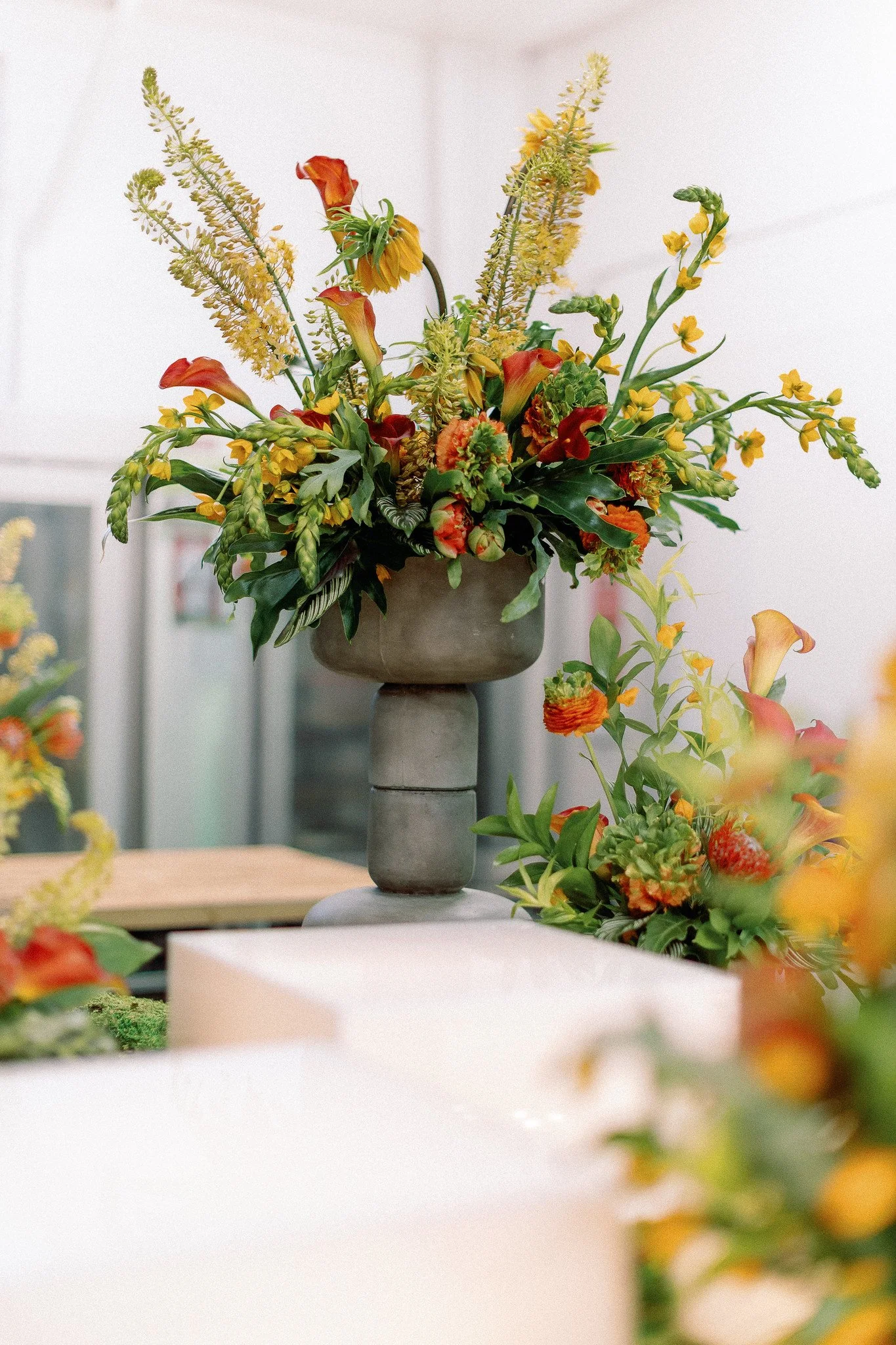 A colorful flower arrangement in a gray vase on a stand, featuring orange, yellow, and green flowers with lush green leaves.
