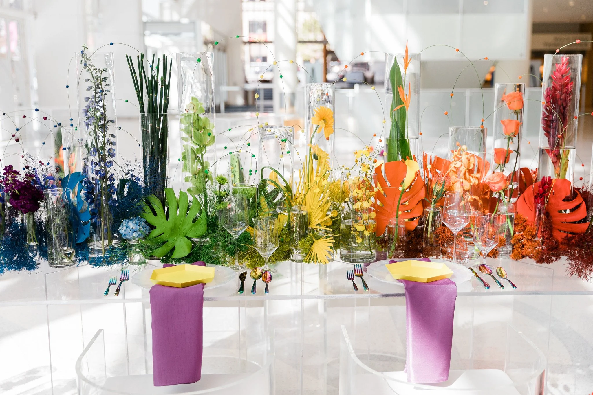 Display of colorful flowers in tall glass vases on a table with purple tablecloths and yellow plates, set in a bright indoor space.