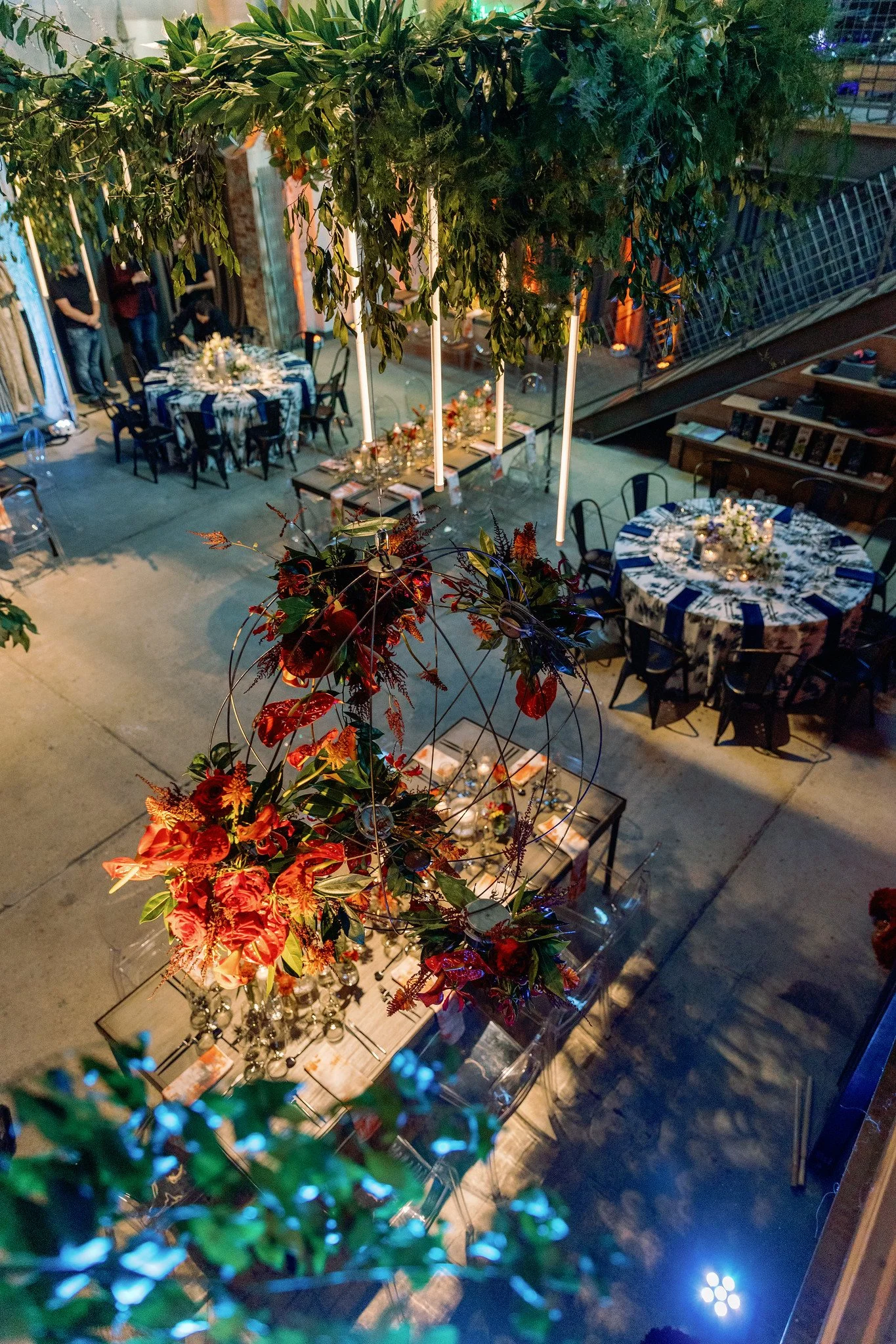 Indoor event space decorated with tables covered in floral tablecloths, floral centerpieces, and candle arrangements, with people in the background. A large green tree or plant is hanging from the ceiling, creating a lush atmosphere. Some people are standing and chatting near the entrance, and there are decorative lighting elements in the space.