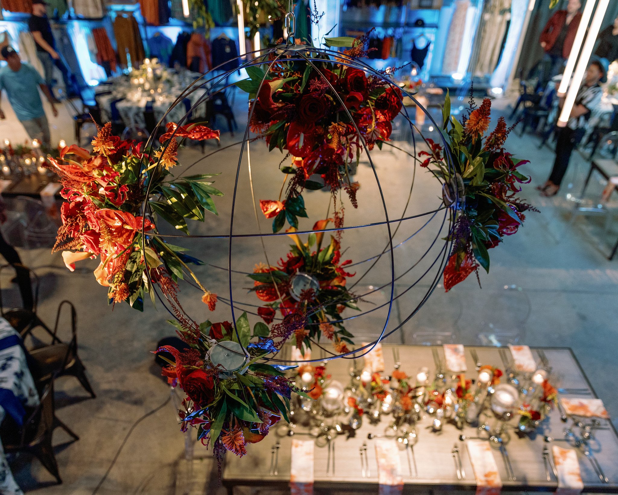 Decorative floral arrangement hanging above a table with blurred guests and decor in the background at an indoor event.