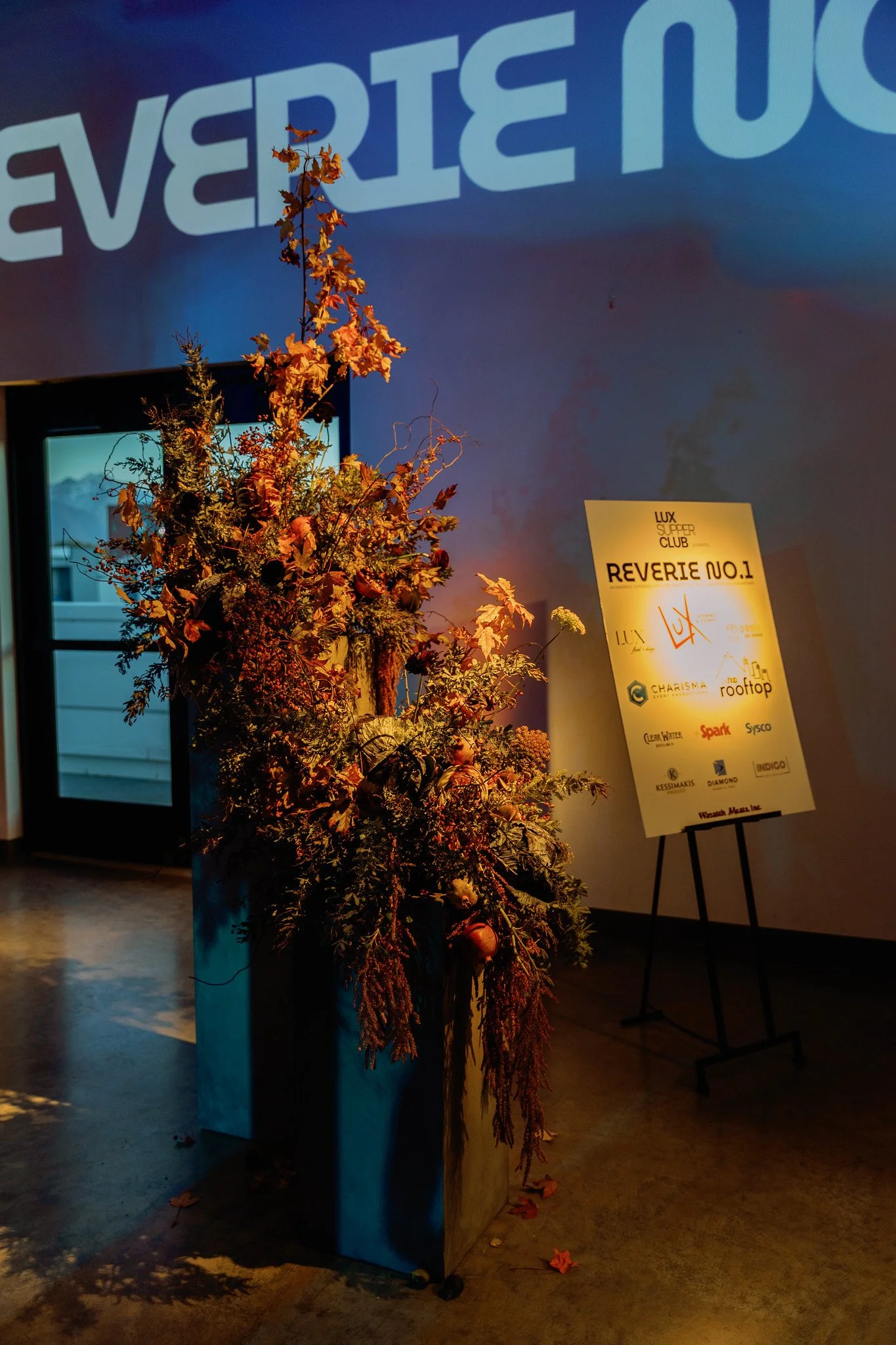 Autumn-themed floral arrangement with dried leaves and berries in a large black vase, in front of a wall with a partially visible illuminated sign and a standing signboard with event details.