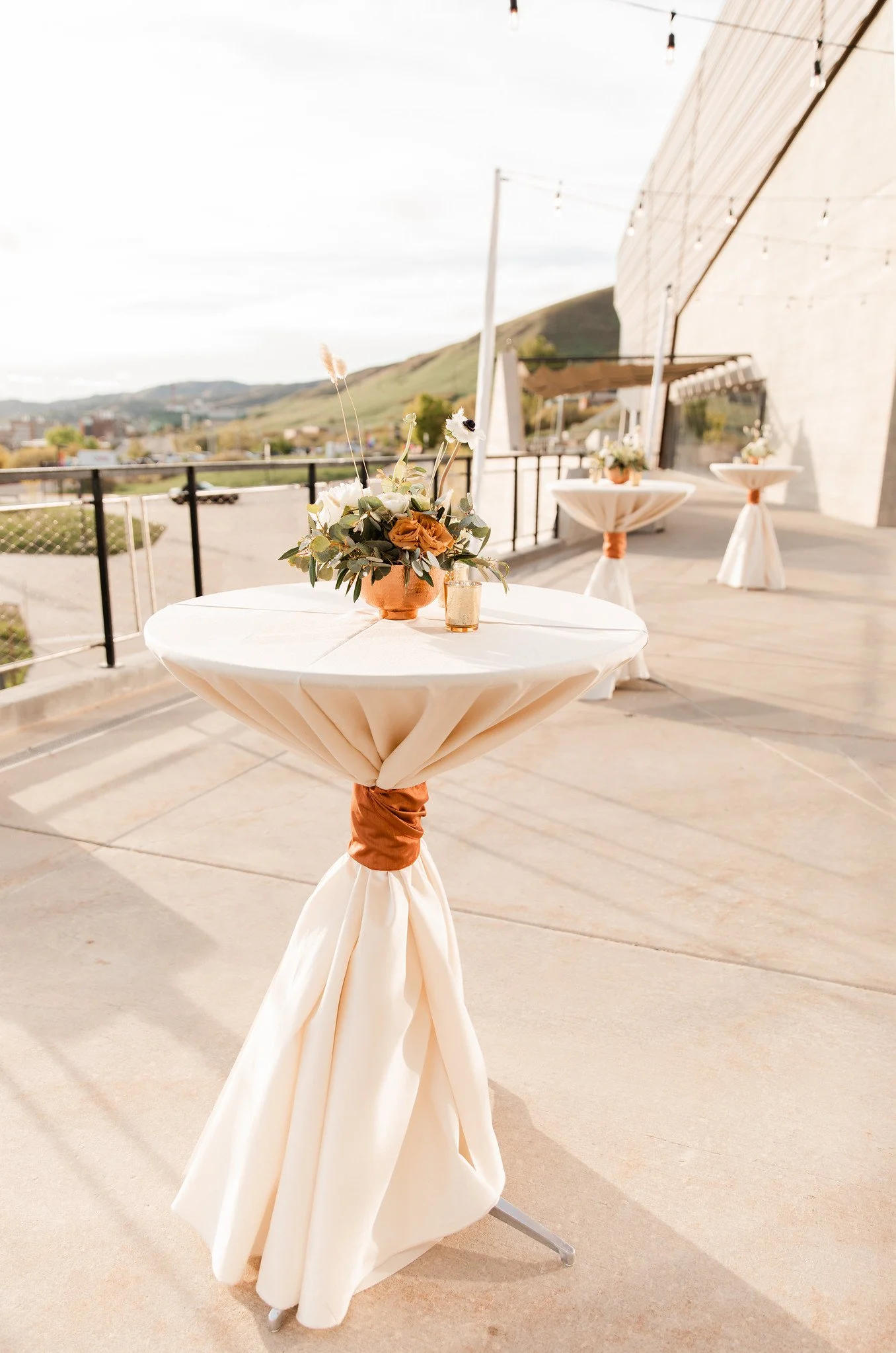 A round table with a beige tablecloth tied with a brown ribbon on a balcony, decorated with a floral centerpiece and a candle holder. More similar tables are in the background, and there is a scenic view of hills and buildings.