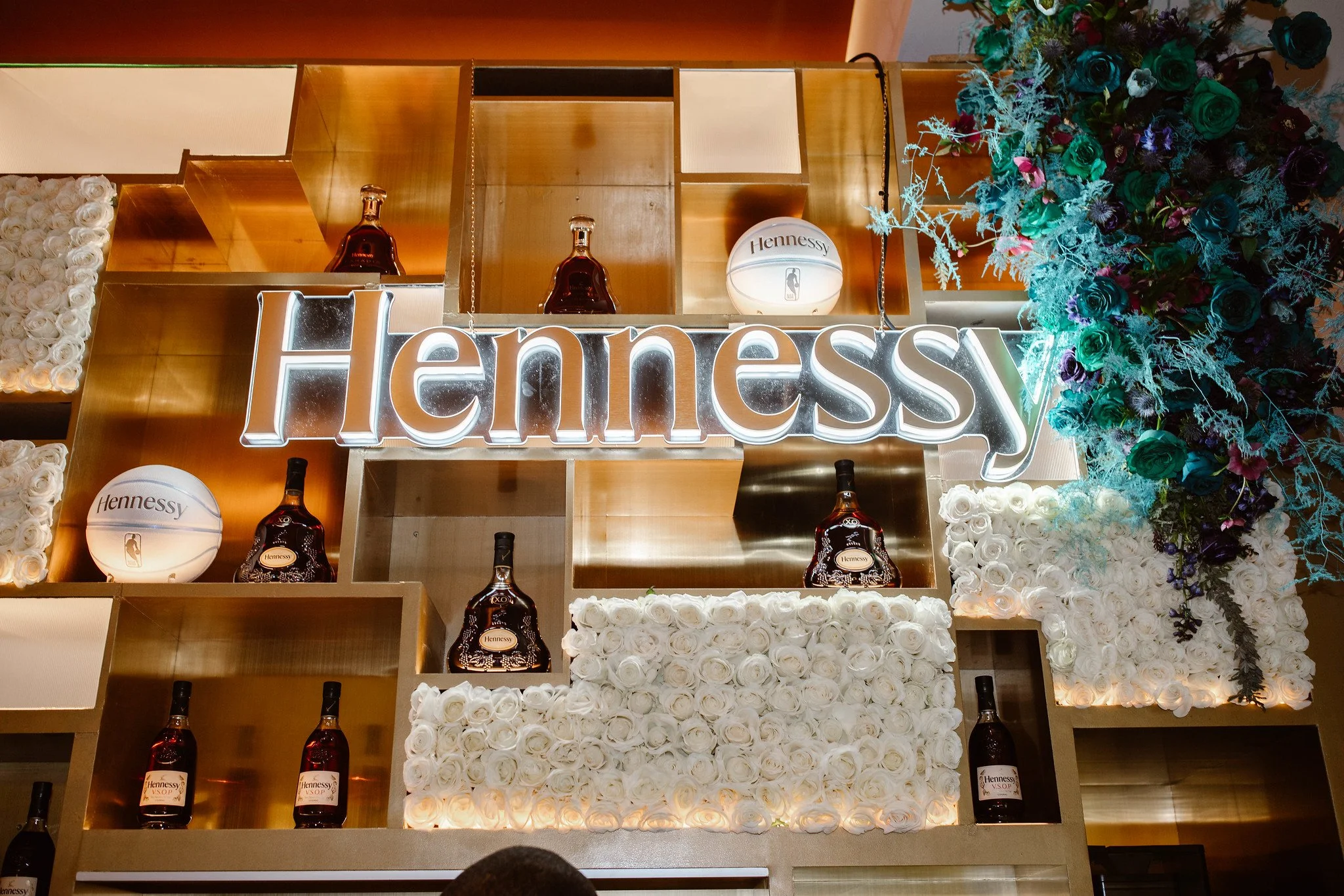 Decorative display with bottles of Hennessy VSOP cognac, a large illuminated Hennessy sign, white roses, a Hennessy basketball, and colorful flowers on top.