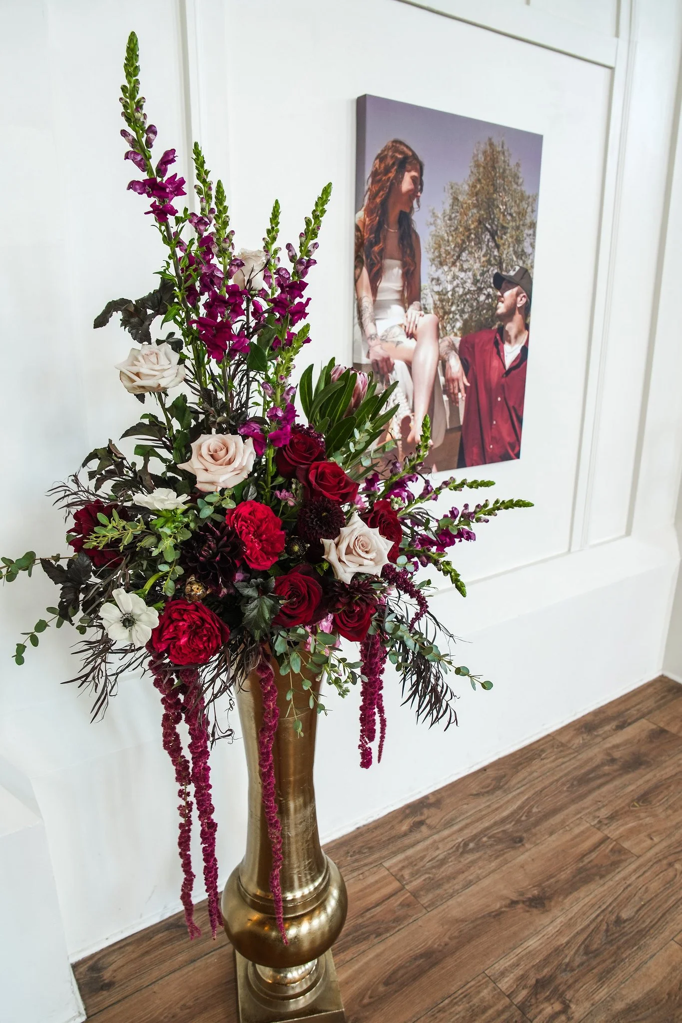 A tall, golden vase with a large, colorful flower arrangement featuring roses, hanging pink amaranthus, and various greenery next to a wall with a framed photo of a woman and man sitting outdoors on a sunny day.