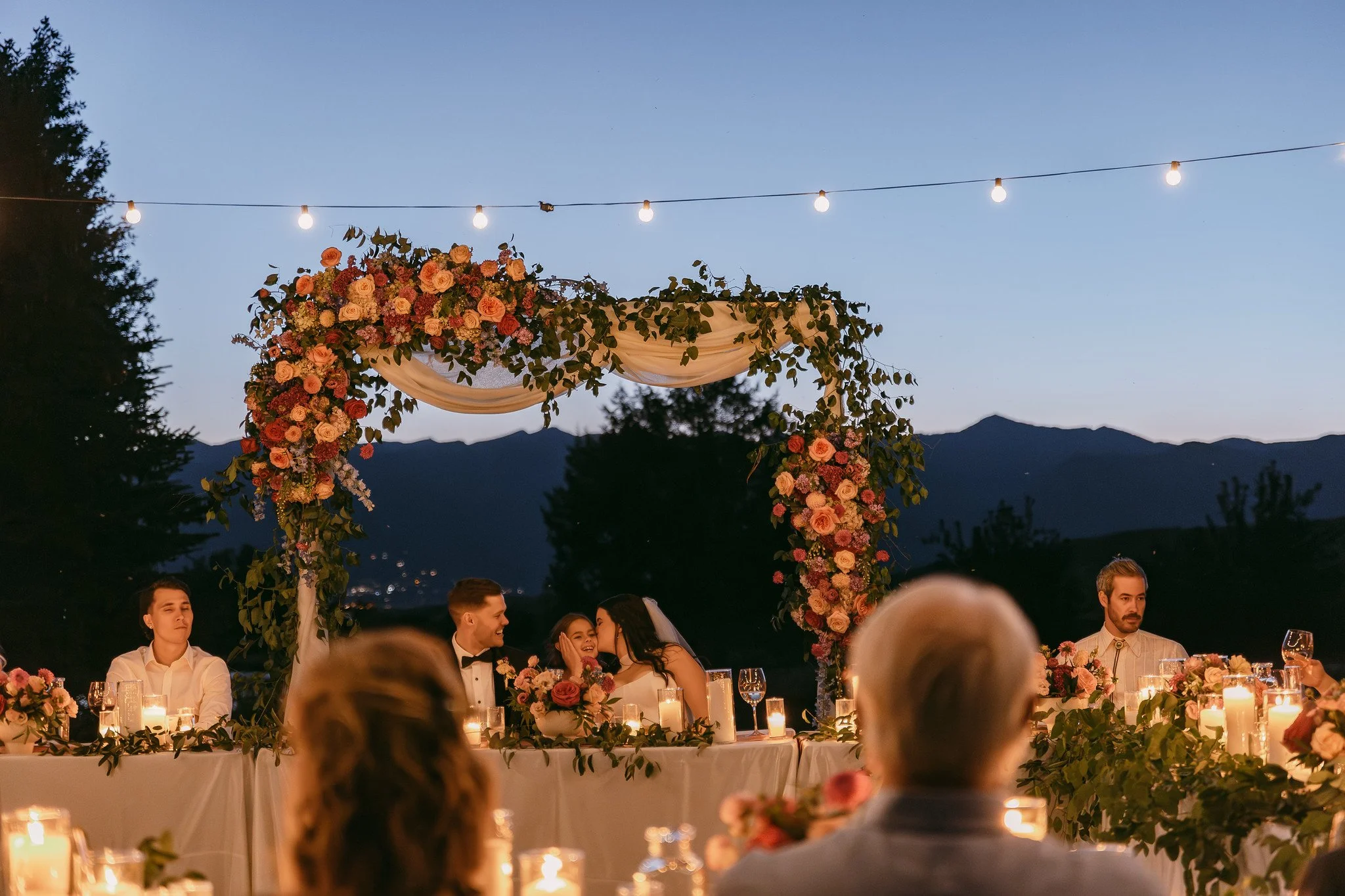 Outdoor wedding reception at dusk with a floral arch and string lights, several guests seated at a long table with candles, mountains in the background.