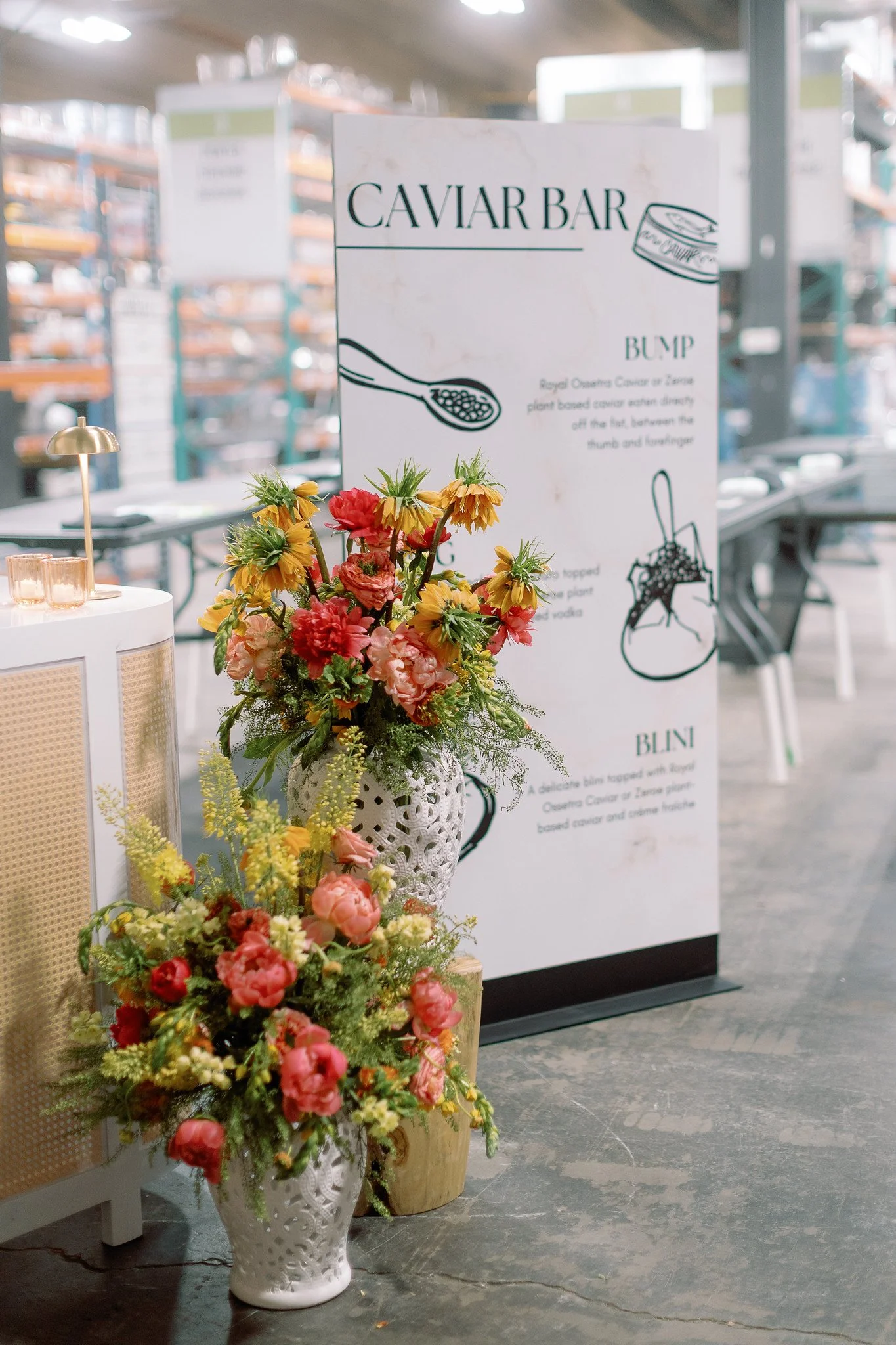 A tall, intricate white ceramic vase filled with pink, red, yellow, and orange flowers, positioned on a concrete floor near a white table. Behind it, there is a white sign with black text and illustrations about caviar, and a blurry background of she