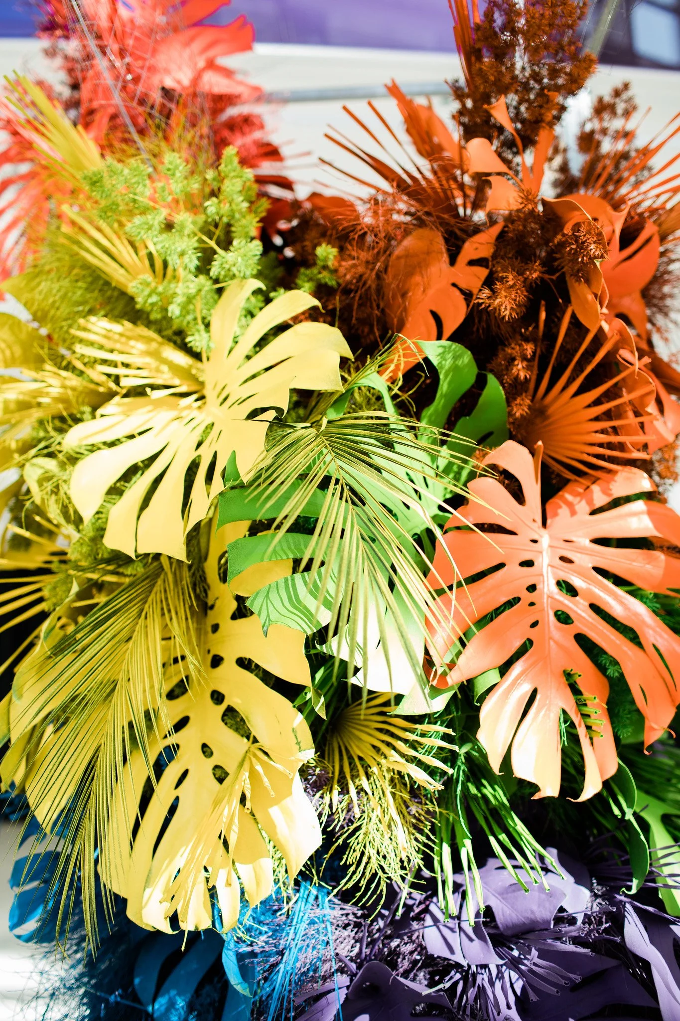 Colorful arrangement of various Caribbean and tropical leaves and plants, including monstera, palm, and fern, with bright and dark hues of yellow, orange, green, purple, and black.