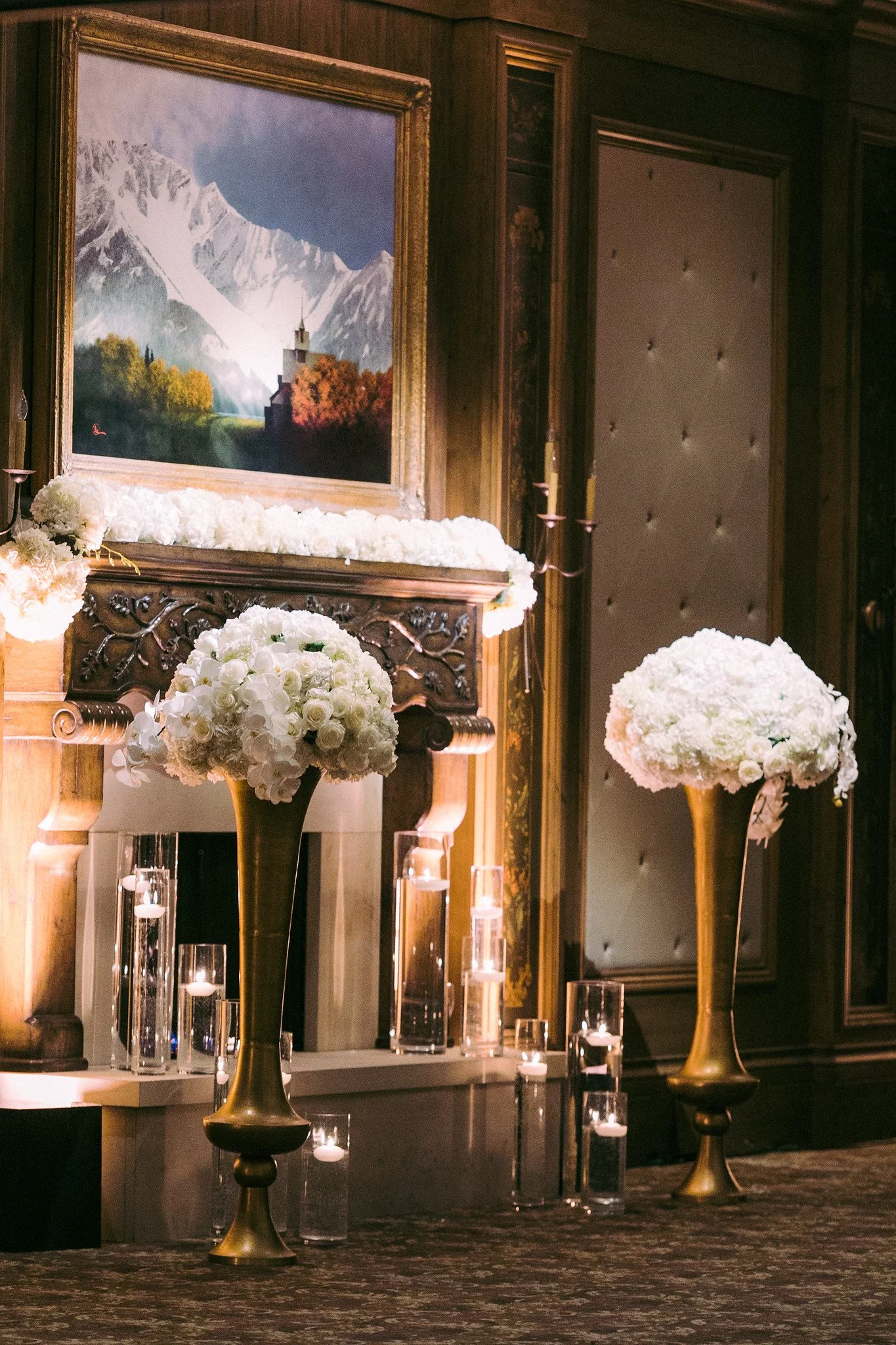 Interior of a room with a mantle decorated with white flowers, gold and black vases, glass candles, and a portrait of a snowy mountain landscape with a church and trees in foreground.
