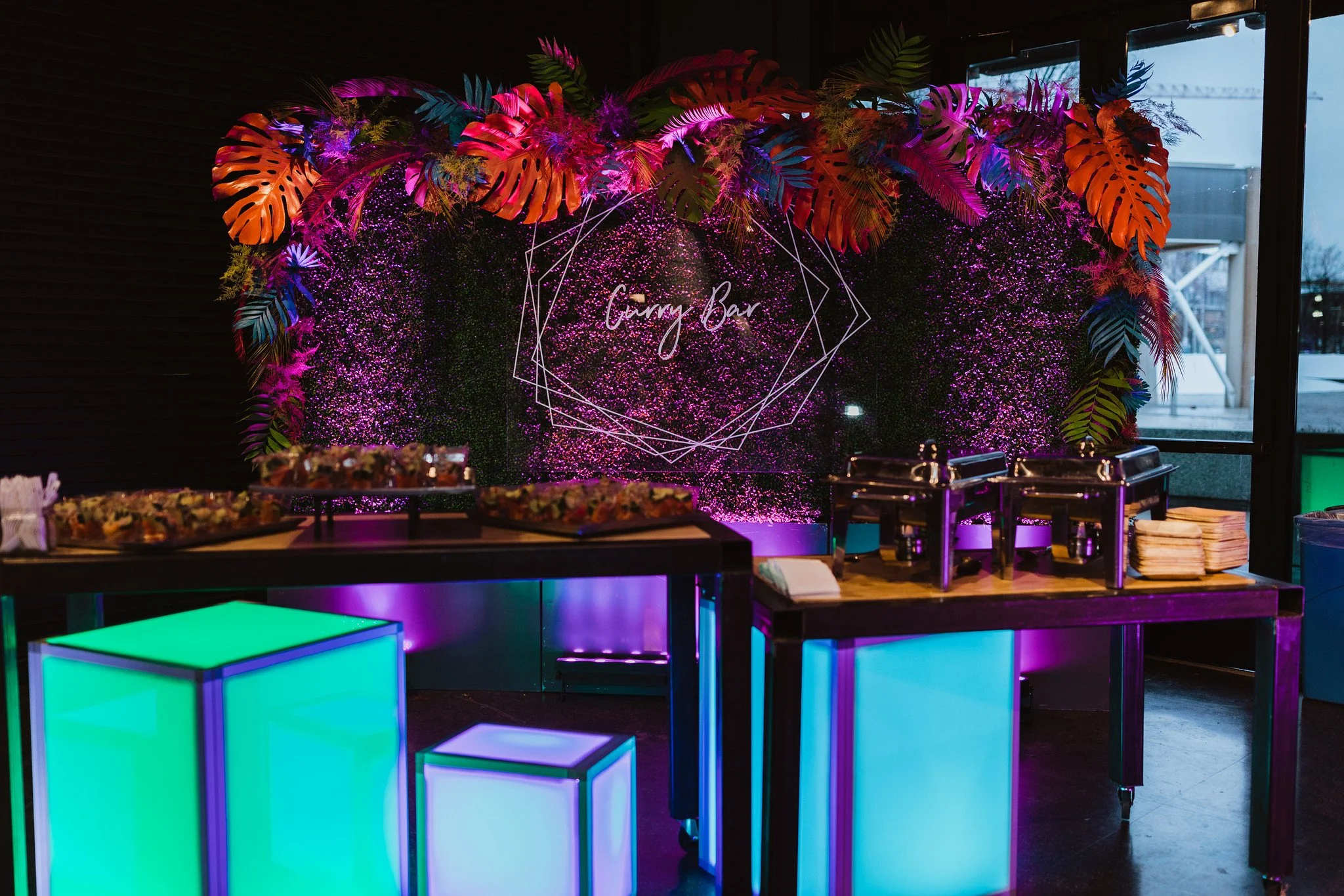 Decorative bar area with colorful tropical leaves, pink and purple lights, and a sign that reads 'Goury Bar' in a geometric frame, with illuminated tables in front.
