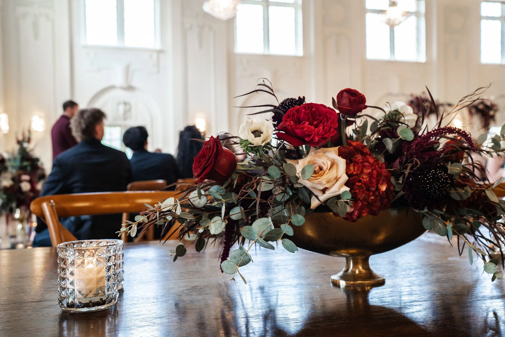 A floral centerpiece with red, white, and dark purple flowers and eucalyptus leaves in a gold vase, placed on a wooden table with a glass candle holder nearby. In the background, people are seated in a bright, elegant room with large windows.