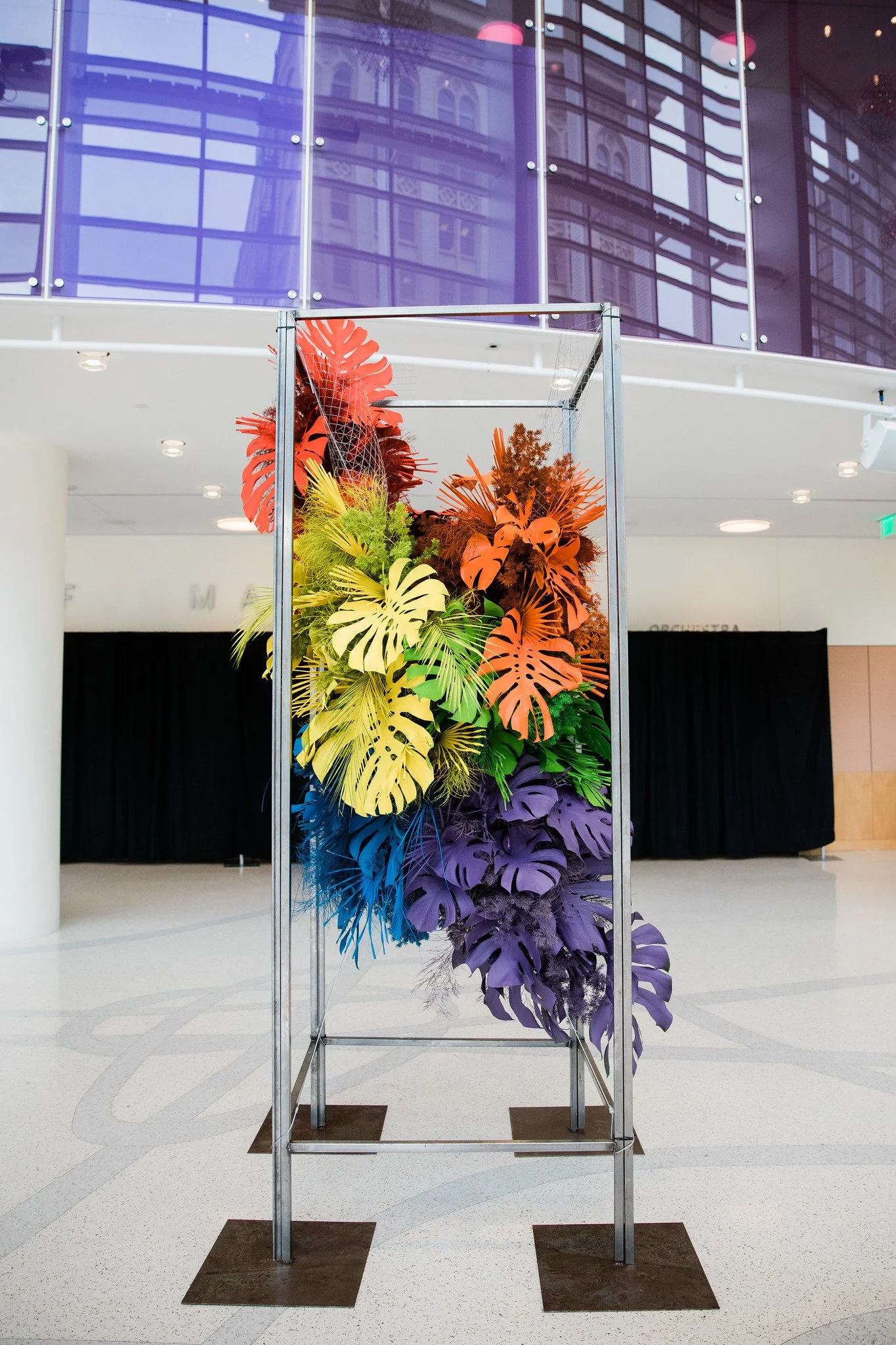 A colorful display of tropical leaves arranged on a metal frame in an indoor space, with a modern glass building with purple-tinted windows reflected on the glass. The floor is white with a curved pattern, and the ceiling has multiple round lights.