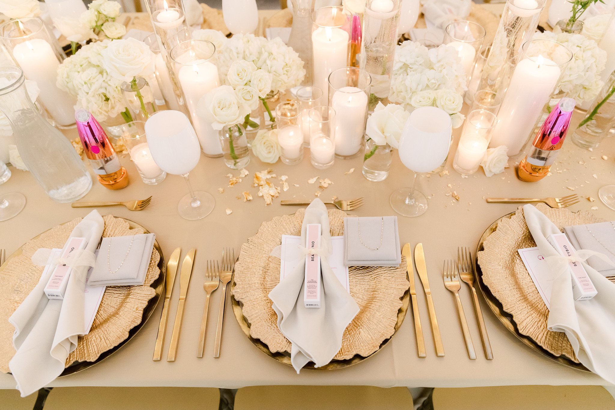 A wedding table decorated with white flowers, candles, and gold utensils, with place cards, napkins, champagne bottles, and glasses.