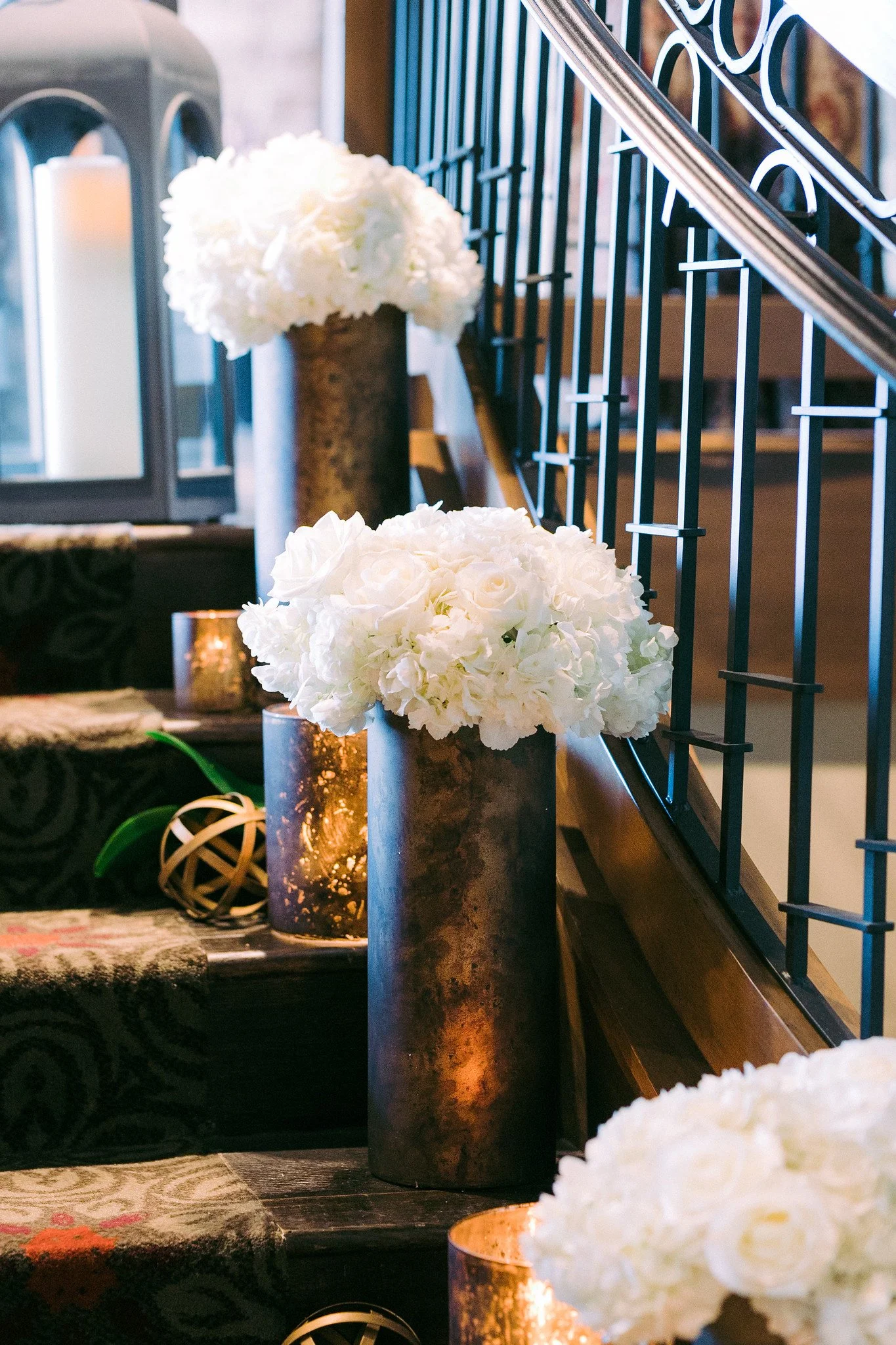 Decorative white floral arrangements in gold vases on a dark wood table beside a staircase with black railings.