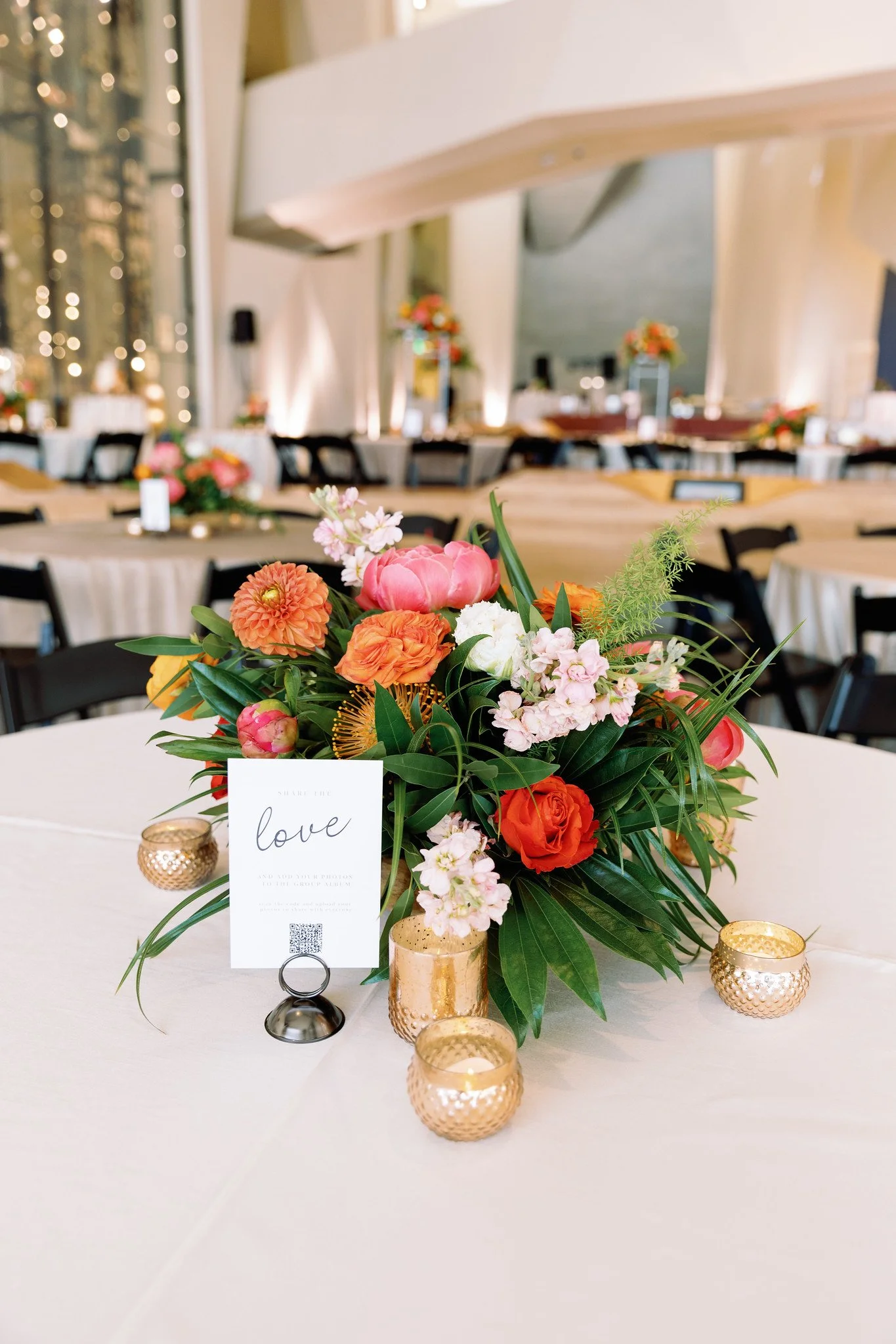 A floral centerpiece with pink, orange, and white flowers on a white table at a decorated event space. Gold candles surround the arrangement, with a placeholder that says "share the love" and a QR code.