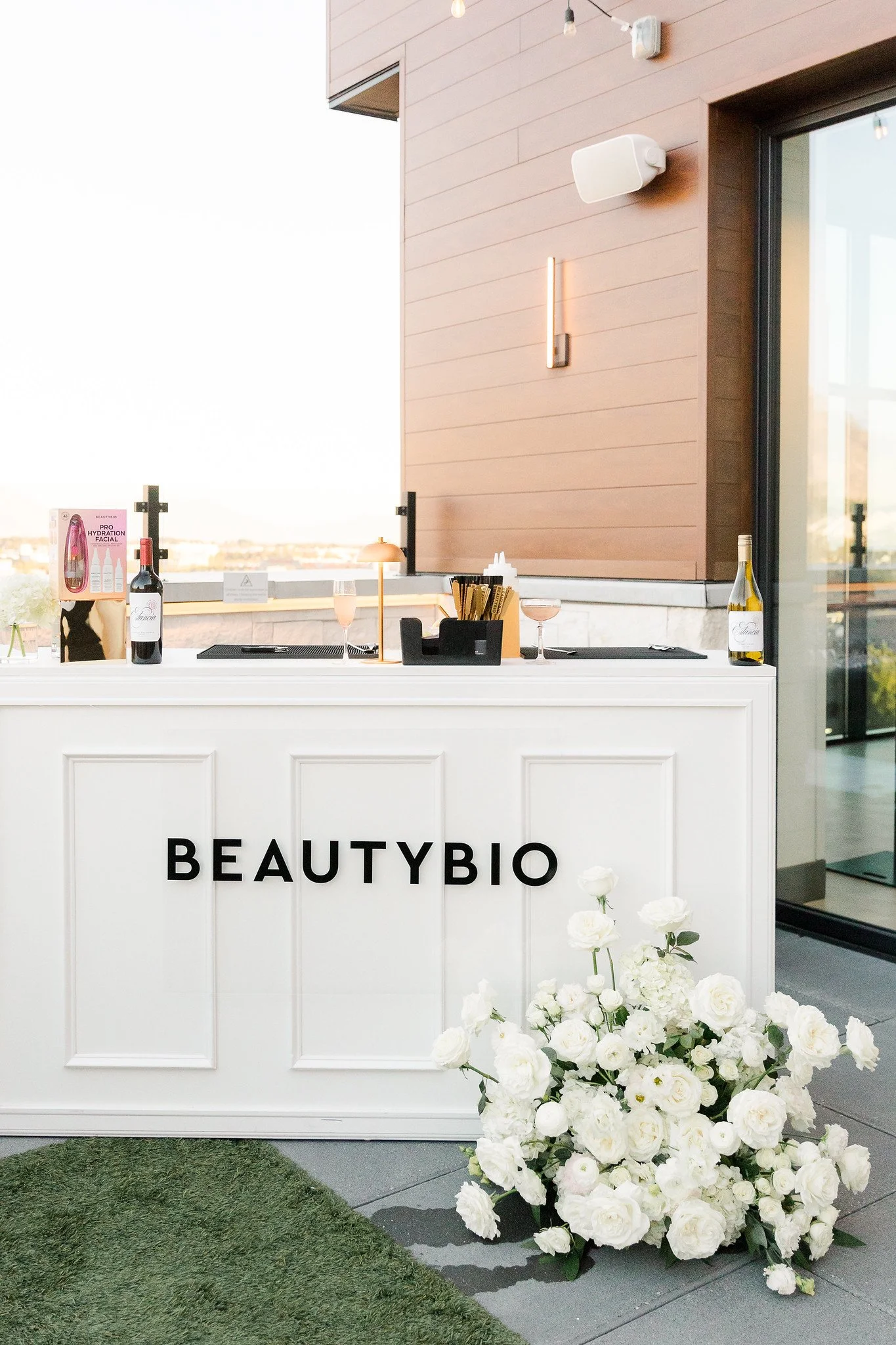 A white reception desk with the words 'BEAUTYBIO' on it, decorated with a large white flower arrangement at the front, and items like bottles, glasses, and skincare products on top, set against a background of a building with a pinkish exterior wall 