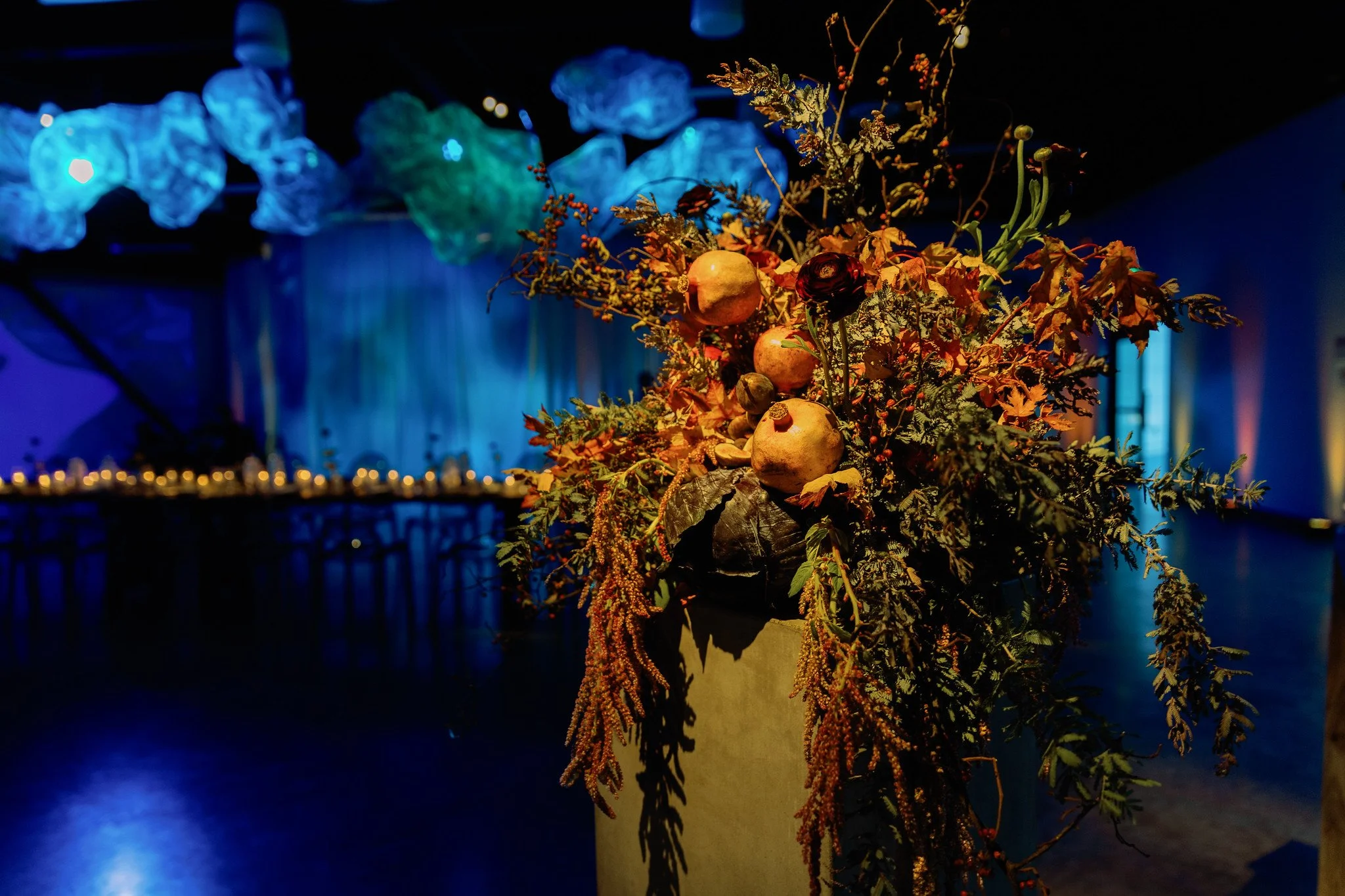 A floral arrangement with dried and fresh flowers and fruits on a pedestal, illuminated by colorful, dim lighting in a dark setting.