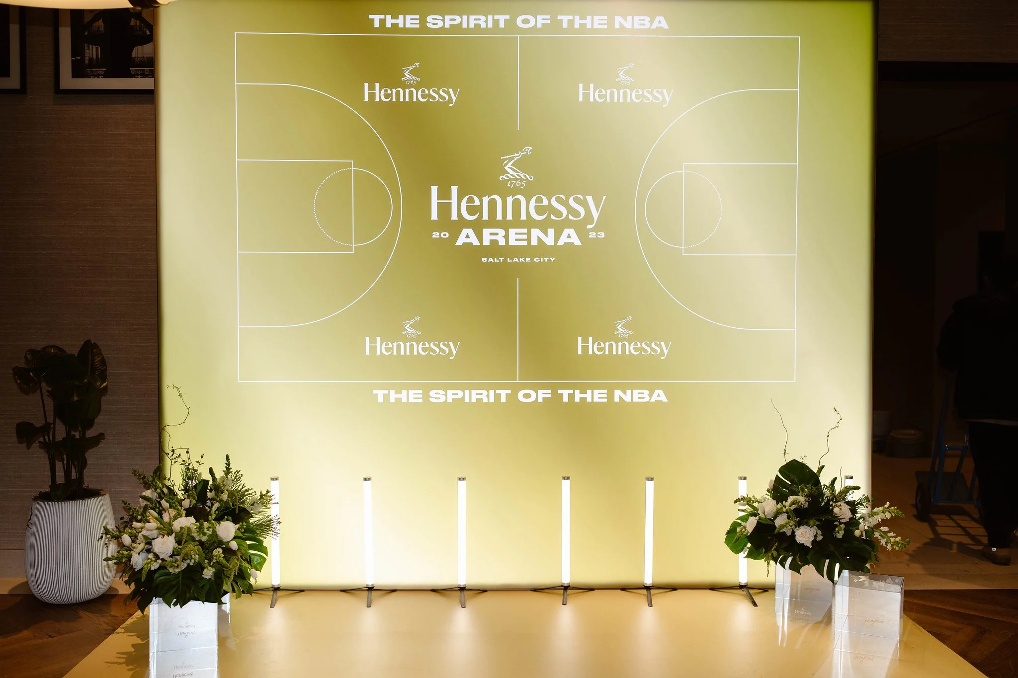 A large yellow backdrop featuring a basketball court layout with the phrase 'THE SPIRIT OF THE NBA' and 'Hennessy' repeated multiple times, located in Salt Lake City, with flower arrangements on either side.