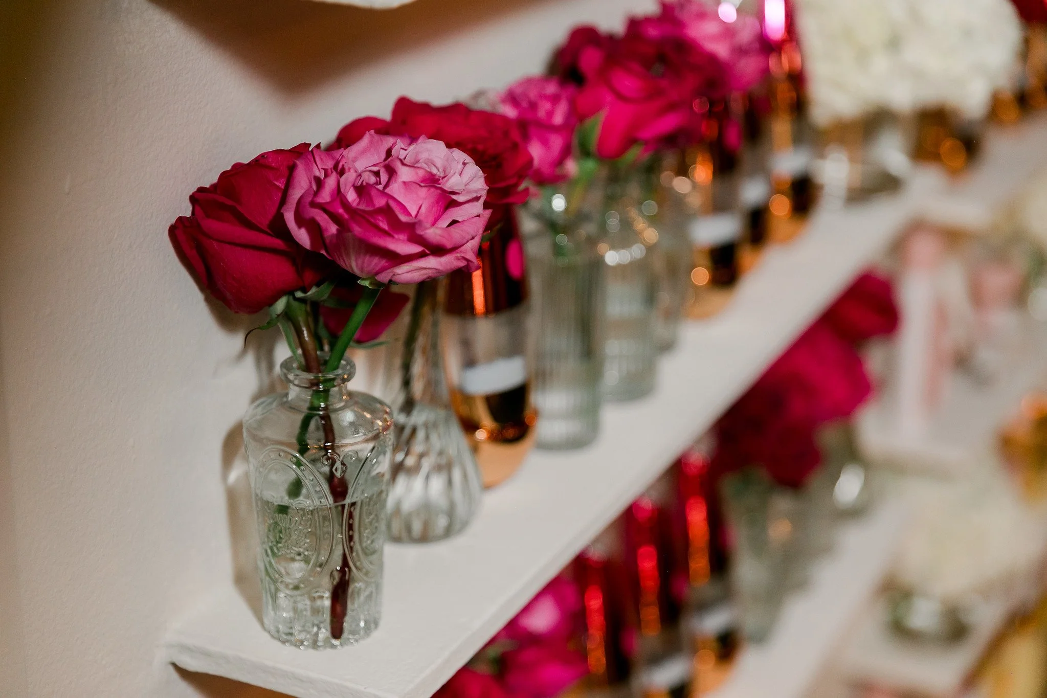A row of small glass vases filled with pink and red roses arranged on a white shelf.