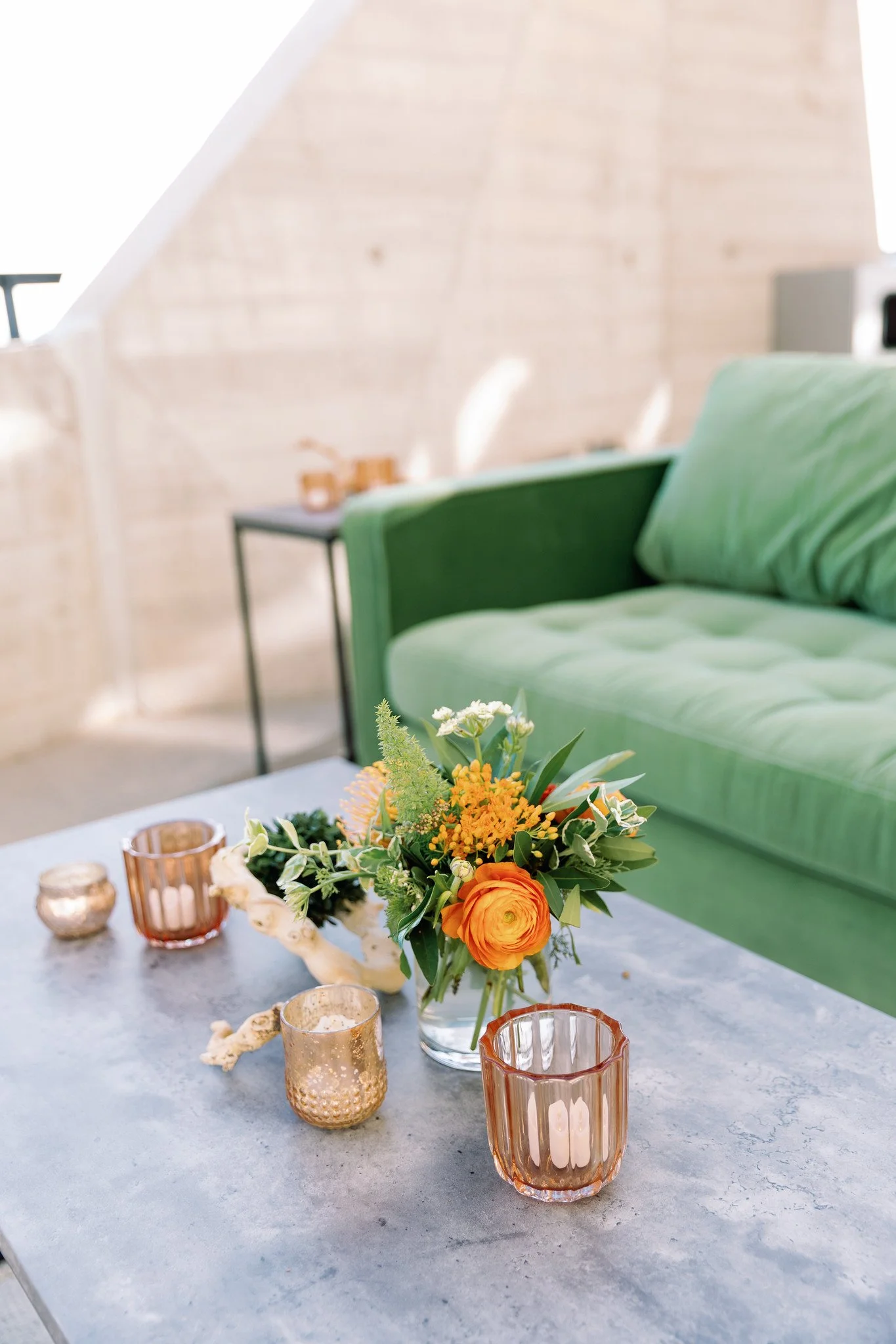 A floral centerpiece with orange roses, yellow and green flowers, and greenery on a gray table in a bright room with a green sofa and a wooden wall.