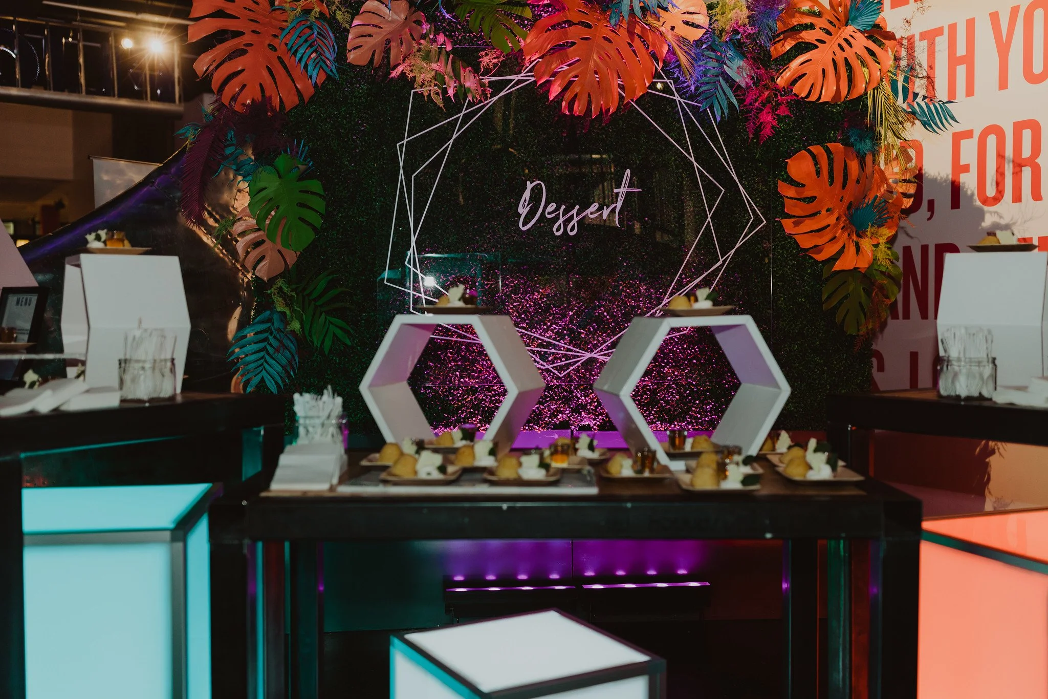 Decorative dessert table with a large green foliage backdrop, pink and orange tropical leaves, geometric white shelves with desserts, and a neon sign that says 'Dessert' at a themed event.