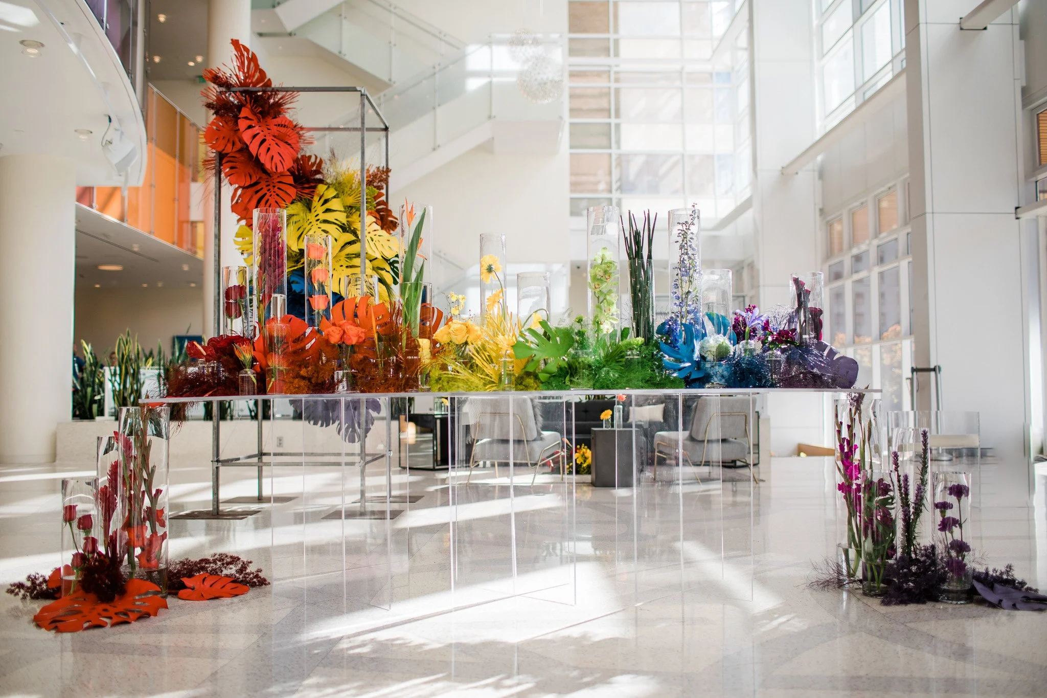 A colorful floral display in glass vases is arranged on a glass table inside a modern, well-lit building with large windows and white walls.