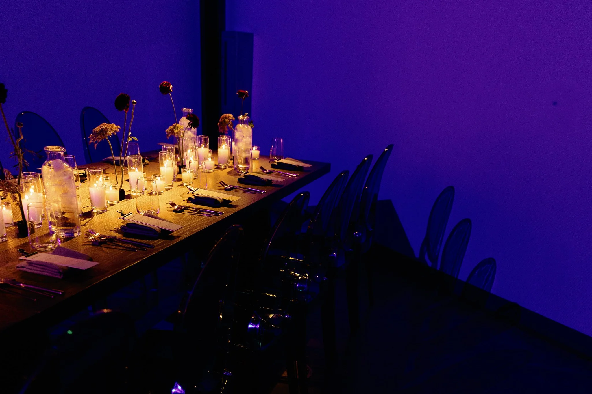 A long dining table decorated with candles and flowers, set for a formal dinner. The room is dimly lit with a dark blue wall background, creating an elegant ambiance.