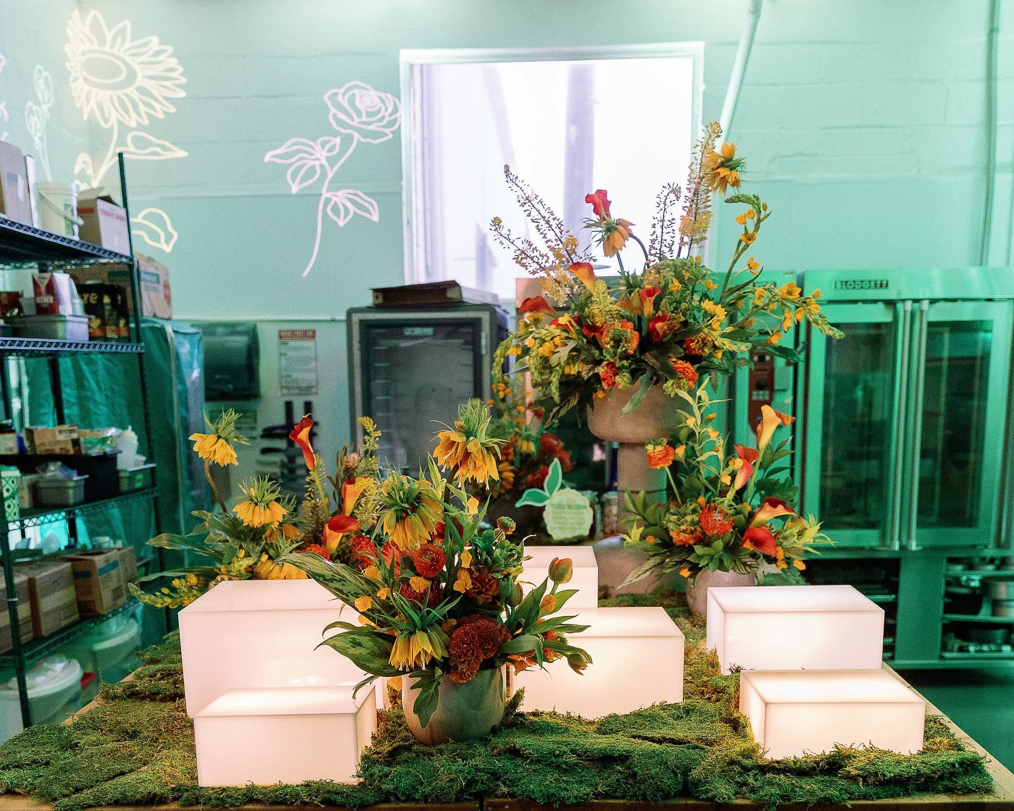 A display of various colorful flowers arranged in a vase on a white platform, with other vases and greenery beneath. The background includes a green wall with white flower and leaf designs, a window, and shelves with storage boxes.