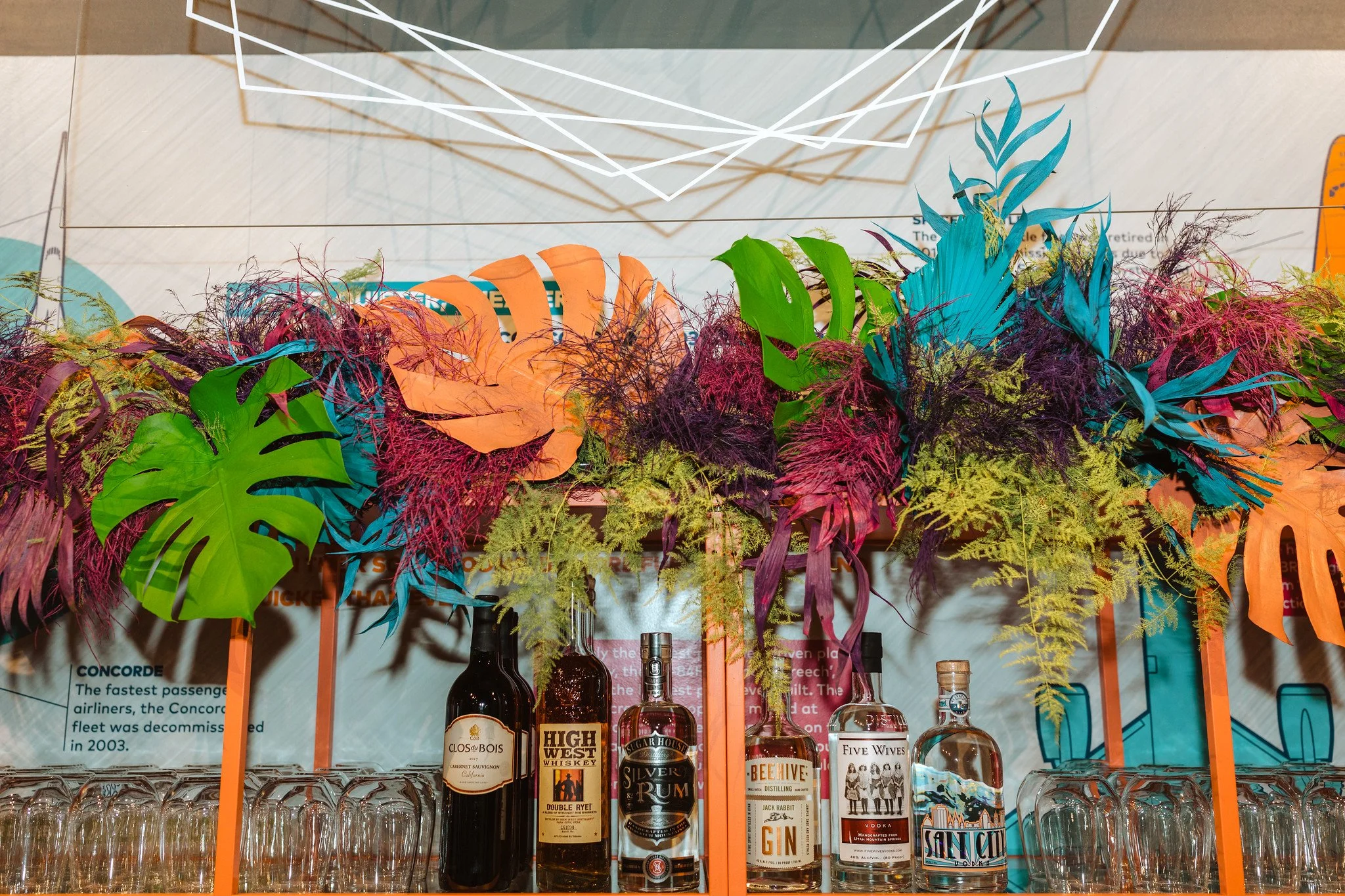 Colorful tropical leaf-themed decorations with liquor bottles and empty glasses in front, displayed on a bar or shelf.