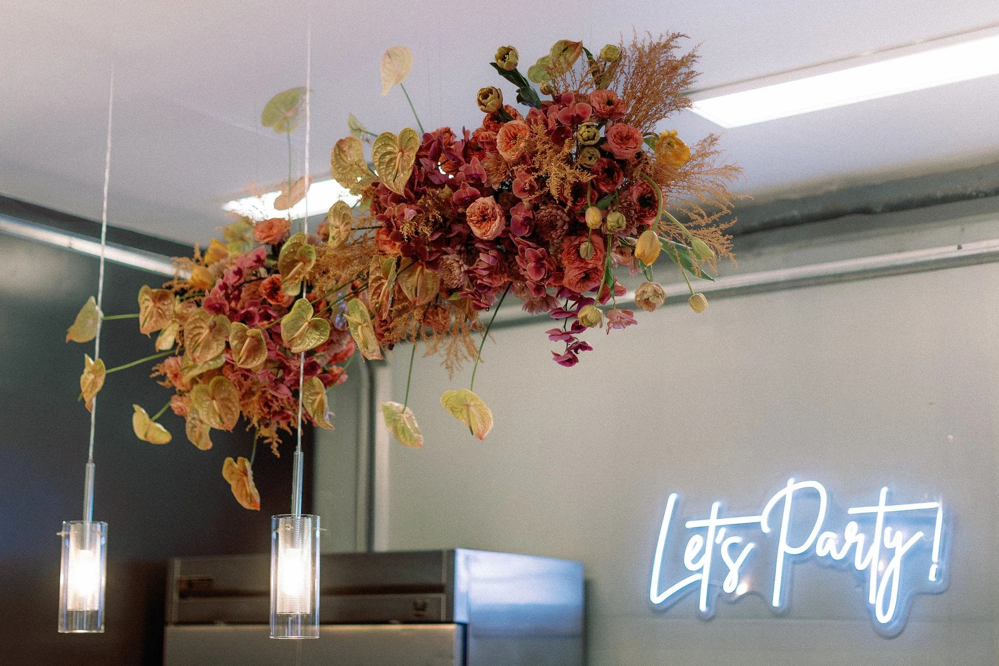 A floral arrangement with pink, orange, and green flowers and foliage hanging from the ceiling in front of a neon sign that says 'Let's Party'