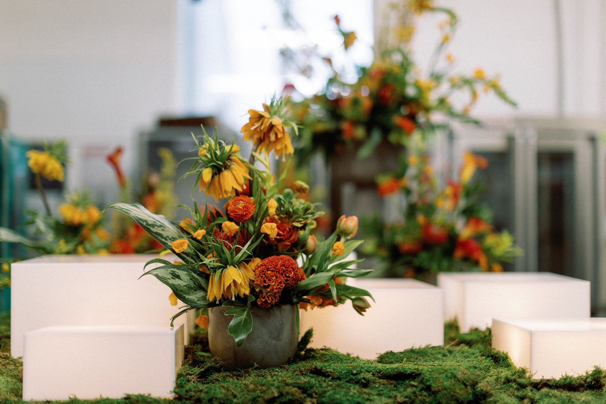 A floral arrangement of colorful flowers including yellow, orange, and red blooms displayed in a gray pot on a white pedestal in an indoor art gallery setting.