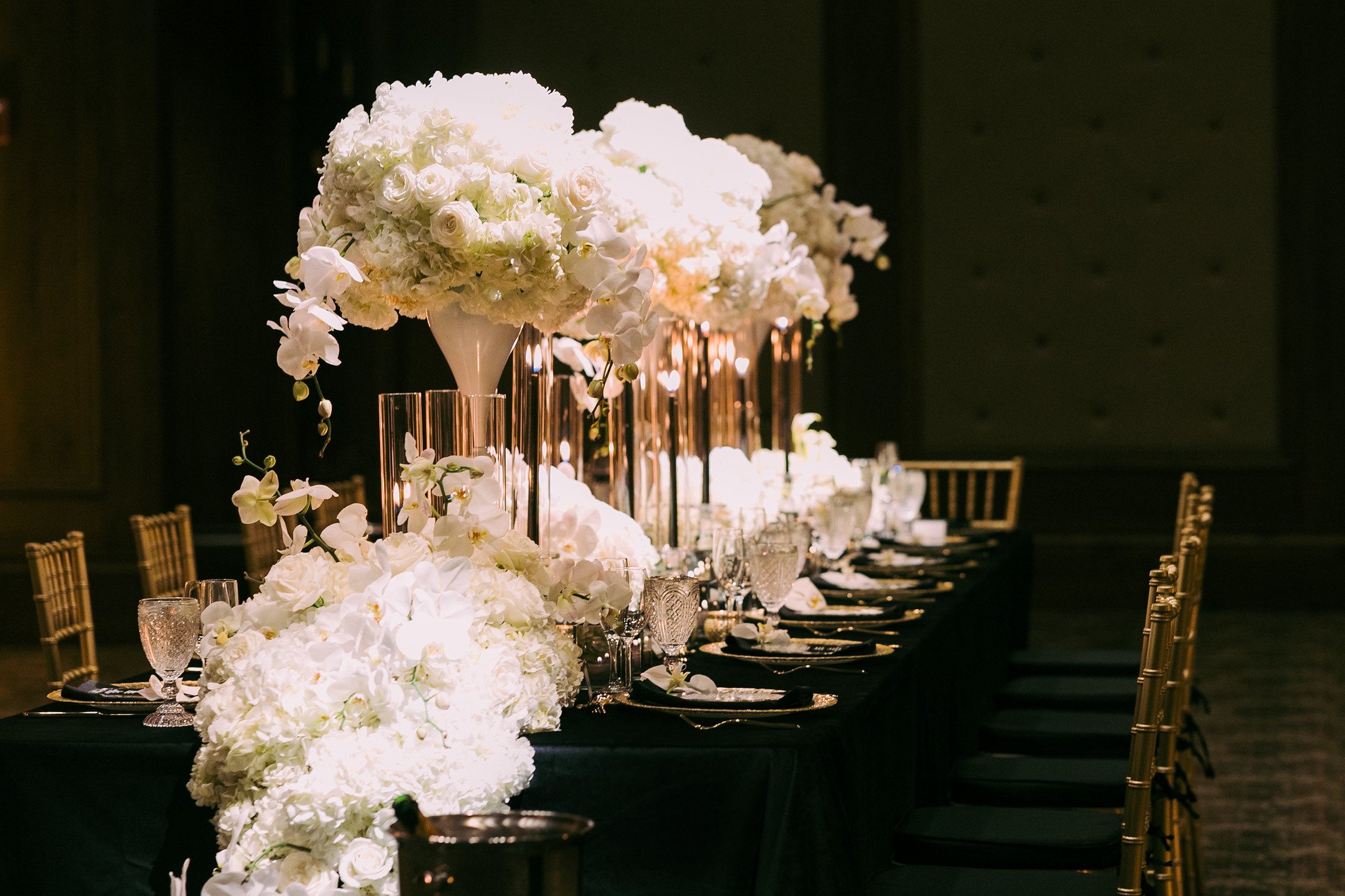 Elegant table setting with tall floral centerpieces featuring white flowers, glassware, and candles on a black tablecloth, in a dimly lit room.