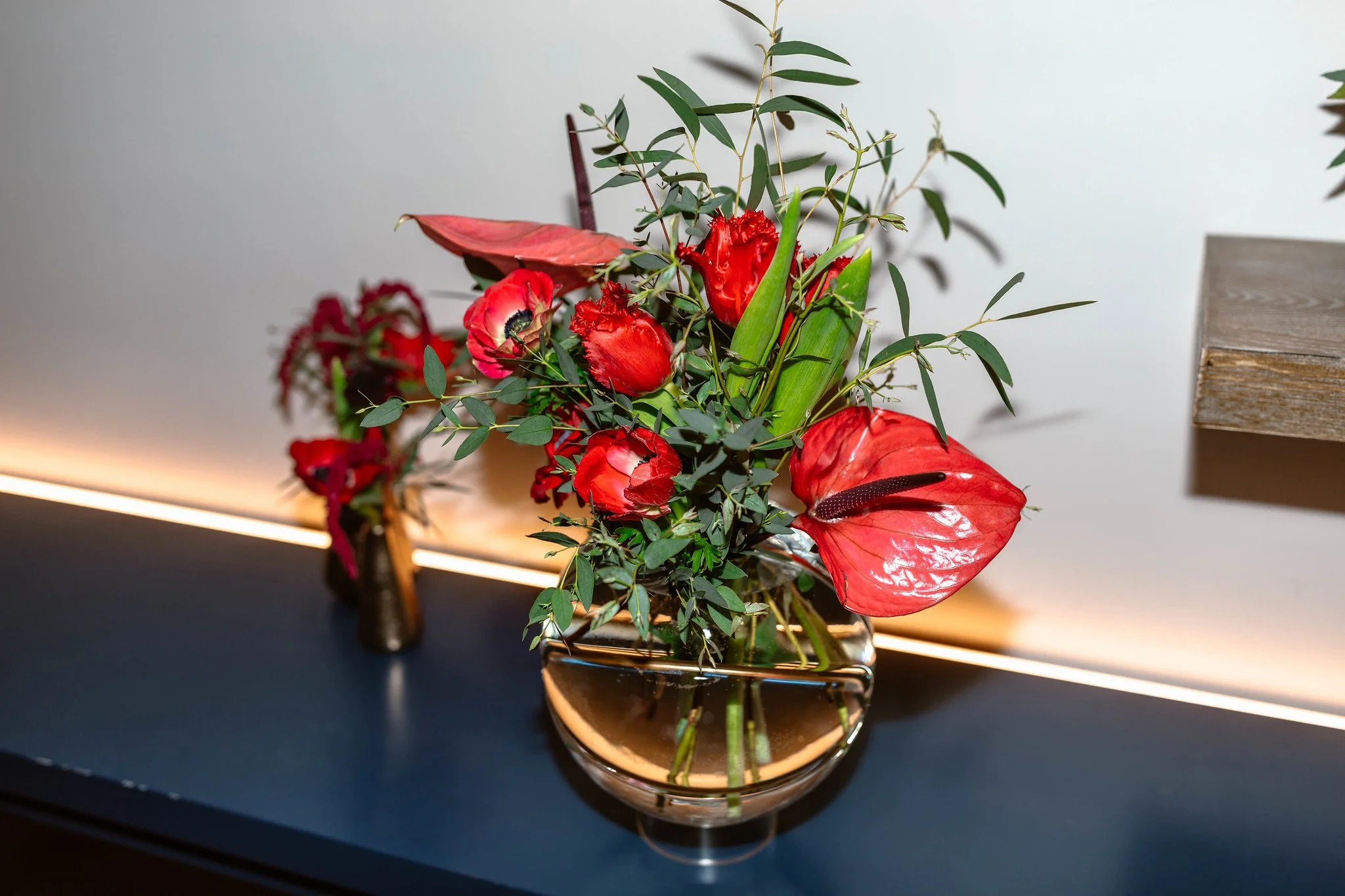 A floral arrangement in a clear glass vase featuring red flowers, green foliage, and a red leaf, placed on a dark surface with a light background.