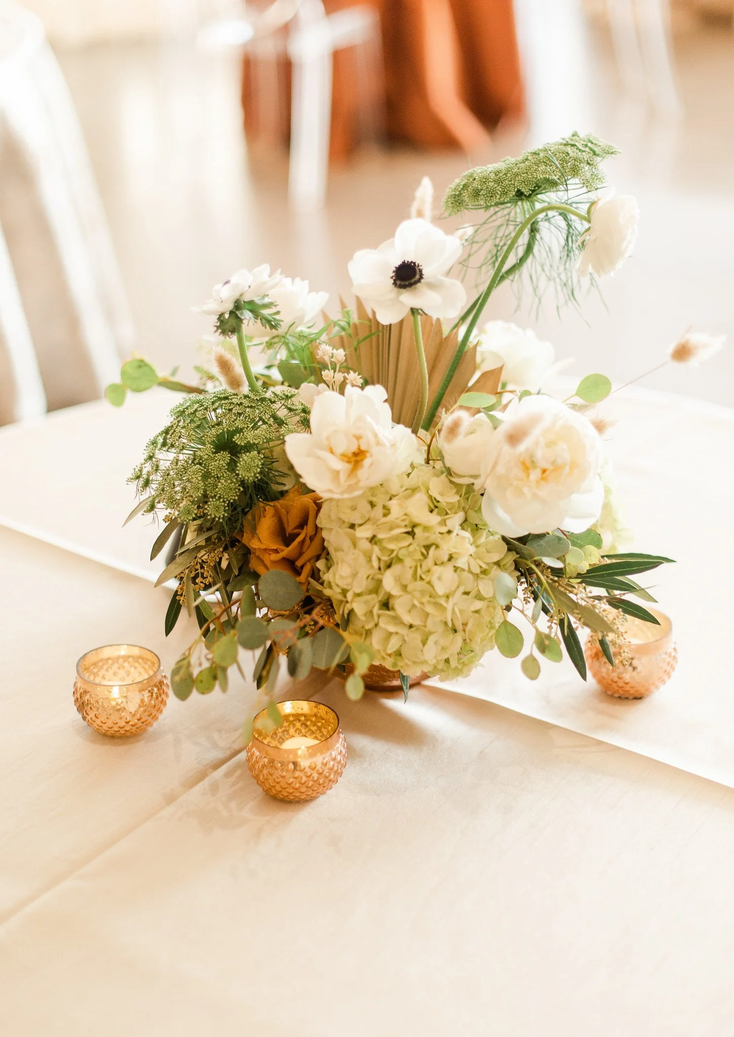 A floral centerpiece with white anemones, green umbels, hydrangeas, and eucalyptus leaves on a table decorated with four small pink glass votive candle holders.