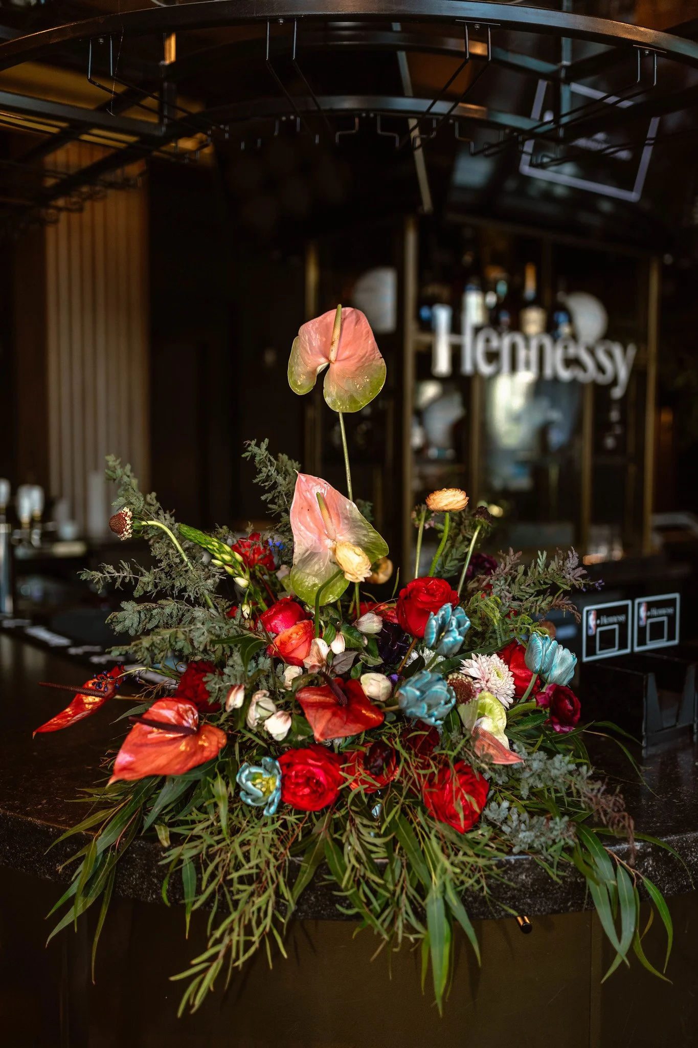 A colorful flower arrangement with red, pink, and blue flowers, green leaves, and anthurium flowers, placed on a black countertop in a bar or restaurant setting, with a Hennessy sign in the background.