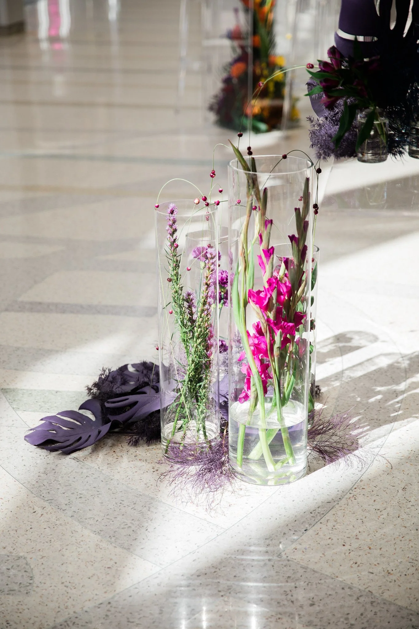 Arrangement of assorted flowers in clear glass vases with purple leaves and decorative purple feathers on white tiled floor.