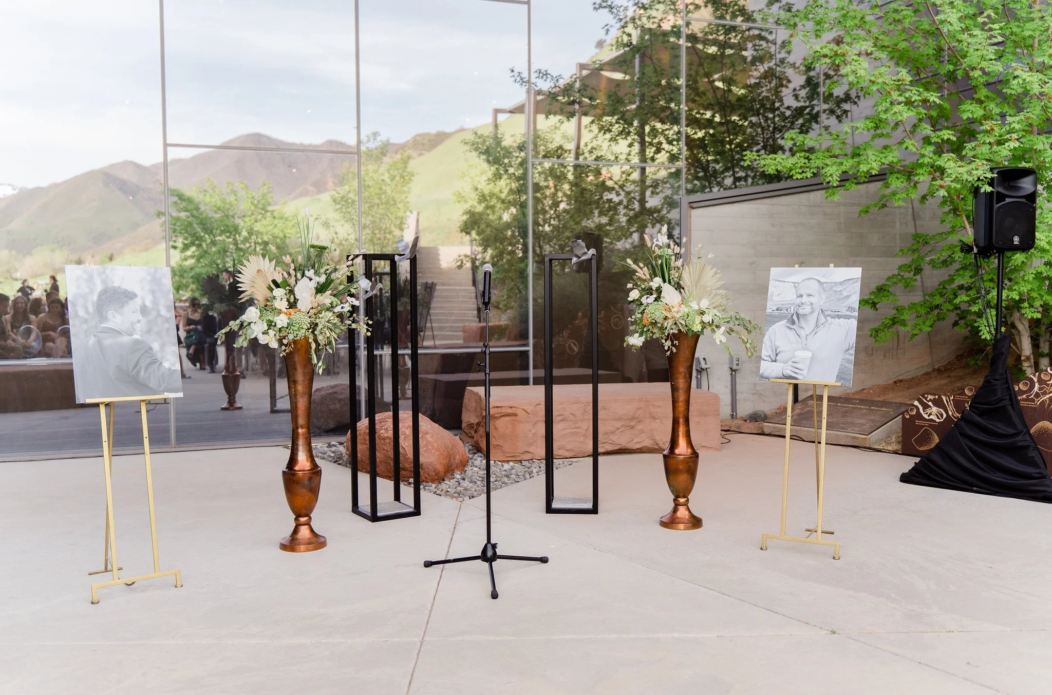 Setup for a memorial or tribute event with two black and white photographs on easels, floral arrangements in tall vases, and a microphone, in front of a glass wall with a view of mountains and greenery outside.