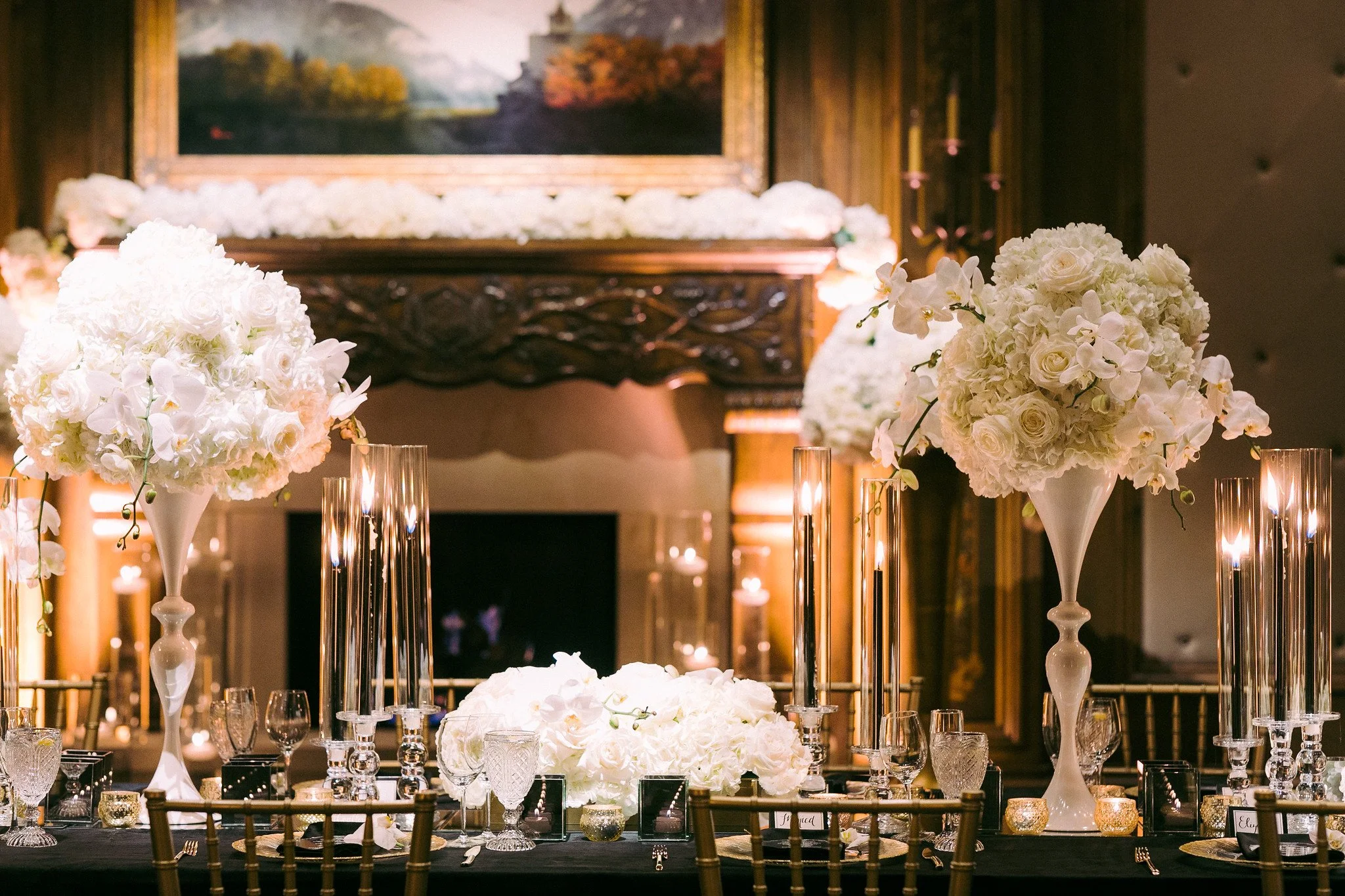 Elegant table setup with large white floral centerpieces, tall white vases, and lit candles in glass holders, set in a dimly lit room with wooden walls and a landscape painting in the background.