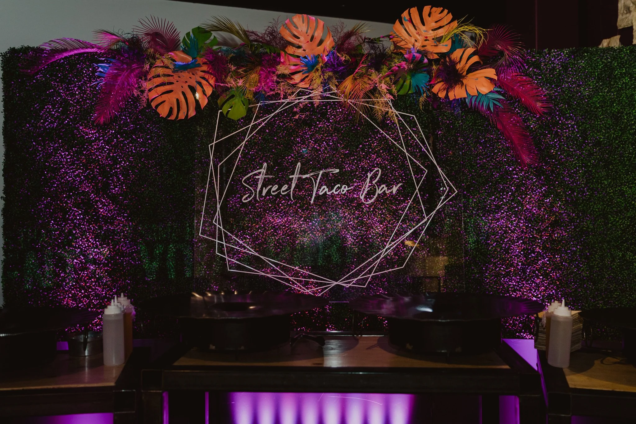 Decorative floral display featuring tropical leaves and flowers above a sign that reads 'Street Taco Bar', illuminated with pink and purple lighting.