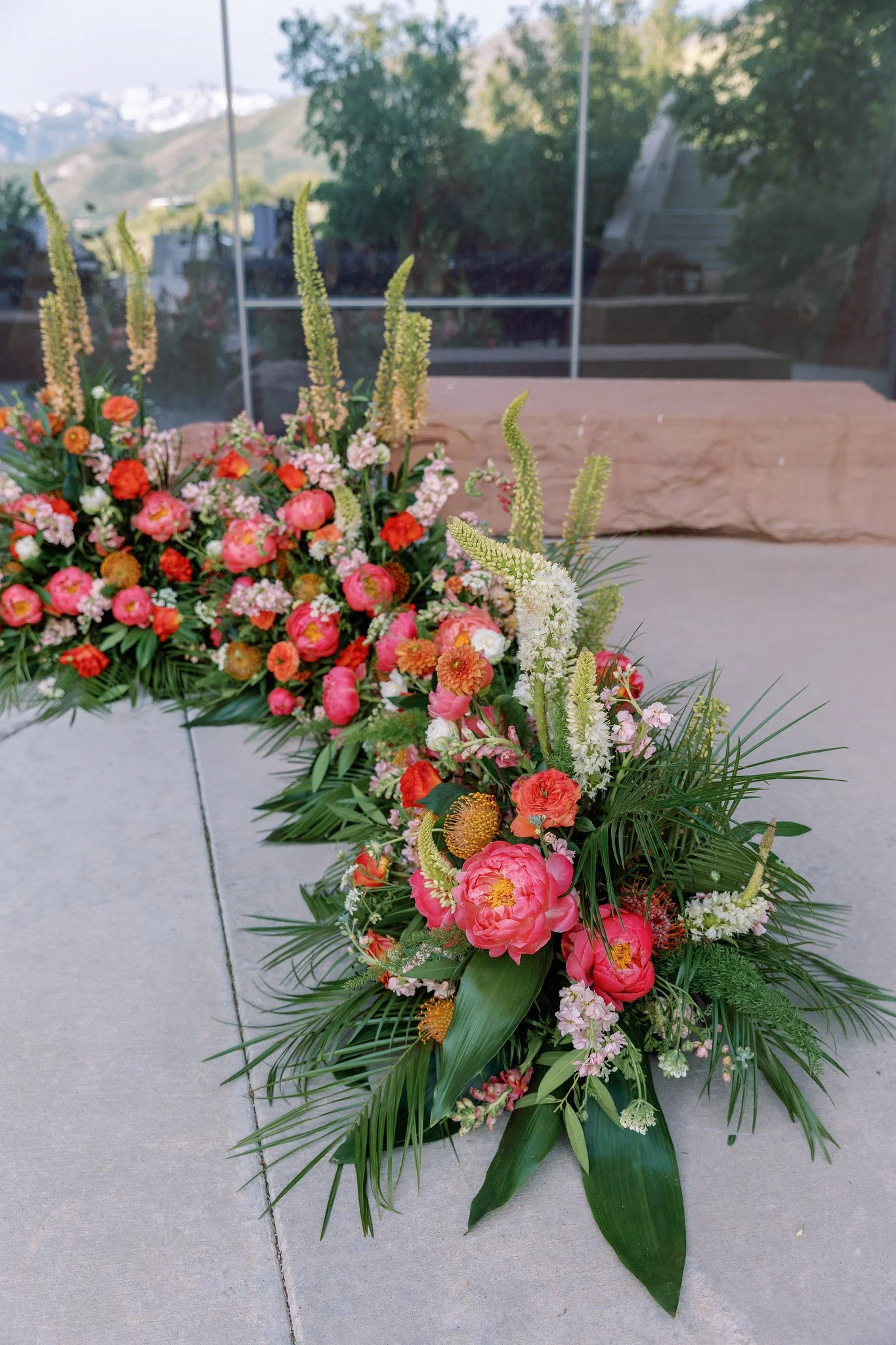 A colorful floral arrangement of pink, orange, and white flowers with green foliage, displayed outdoors near a wooden bench, with a background of trees, mountains, and a glass building.