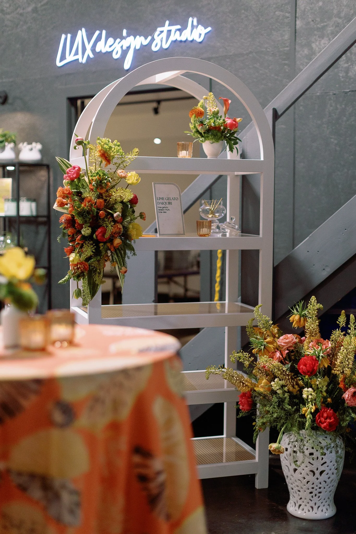 Interior space with floral arrangements, a sign reading 'LAX design studio' in neon, and a decorative white ceramic vase with flowers, set against dark walls and a staircase.