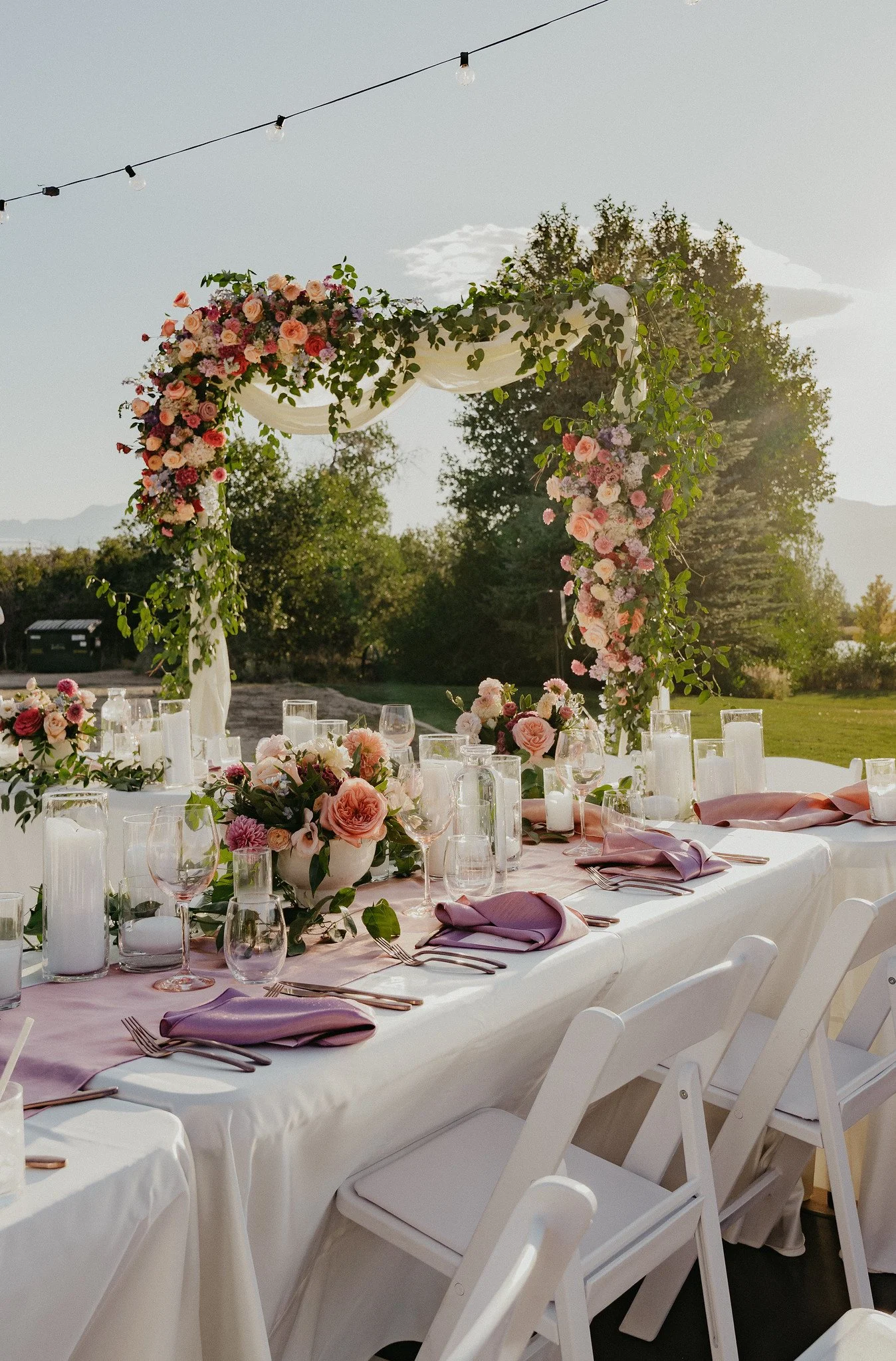 A long outdoor wedding reception table decorated with pink and purple flowers, candles, and wine glasses. A floral and greenery arch is positioned at the end of the table, with string lights hanging above. The scene is set in a grassy area with trees and mountains in the background.