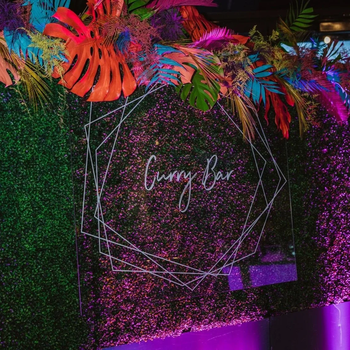 Colorful tropical leaves and purple flowers with a neon sign that reads 'Gummy Bar' surrounded by geometric lines on a green and pink flower wall behind a bar area.