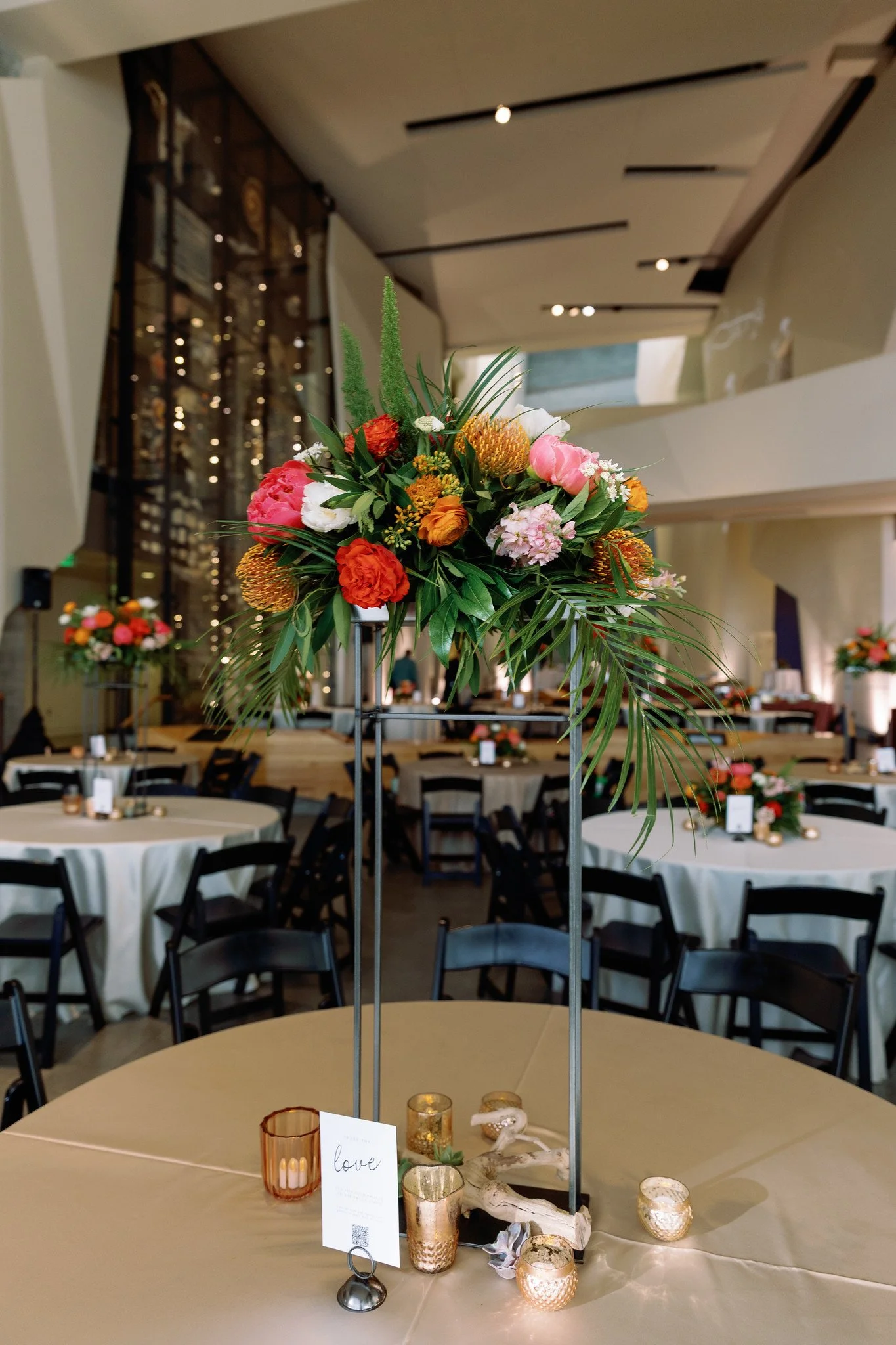 Table centerpiece with a floral arrangement of pink, orange, and white flowers and green foliage, surrounded by candles and decorative items on a beige table in an event space.