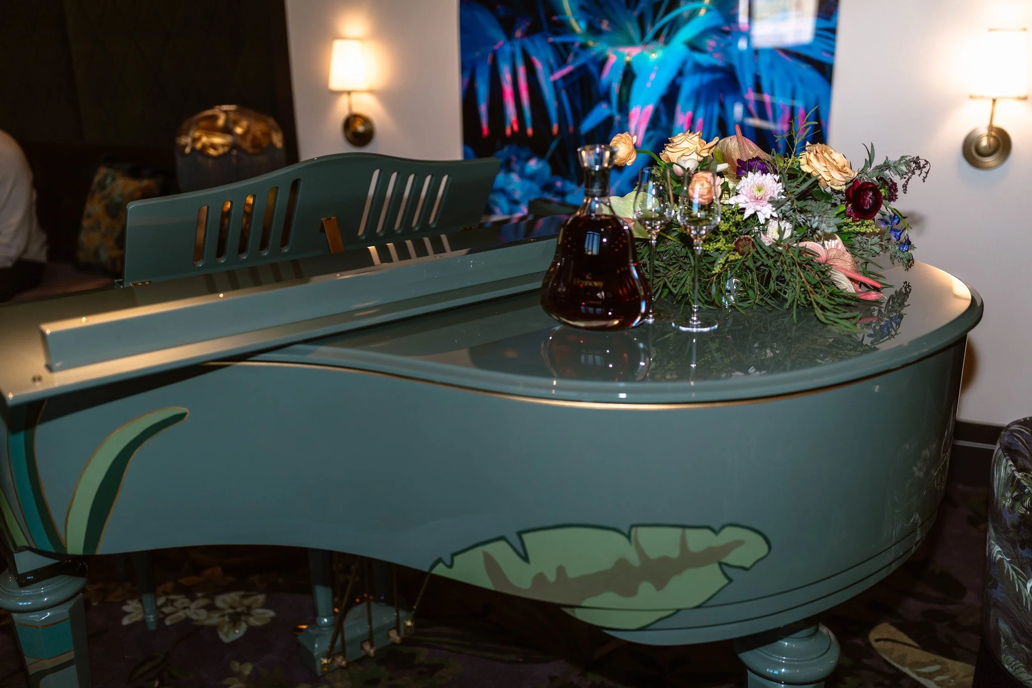 A teal-colored piano decorated with a floral arrangement and two glasses of wine, set in a dimly lit room with wall-mounted lamps and a vibrant blue abstract painting in the background.