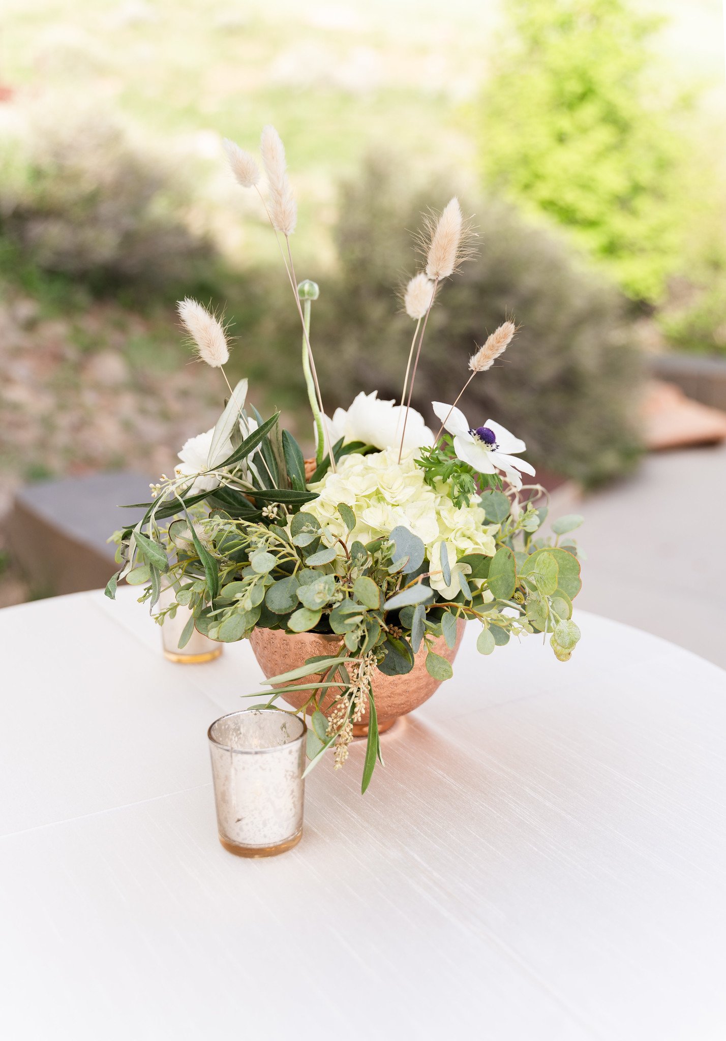 A floral centerpiece in a copper vase containing white flowers, greenery, and tall dried grass-like plants, placed on a white table with a glass candle holder in front.